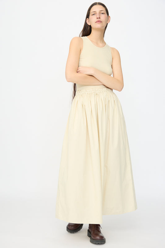 A person stands against a plain background wearing the AVN Tank Dress in Sand, a sleeveless cotton dress with a long gathered skirt, styled with brown lace-up boots.