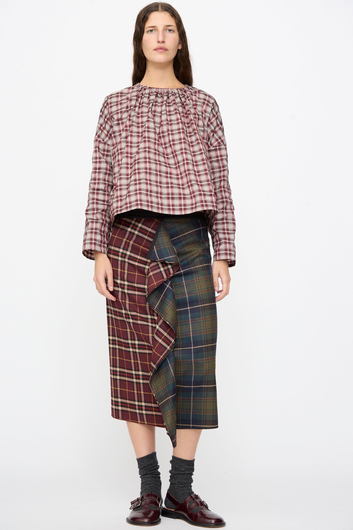 A person wears the Volant Skirt in Bordeaux and Military Green Check, a burgundy and green plaid wool midi skirt, with a ruffled front. Worn with a plaid top and burgundy loafers.