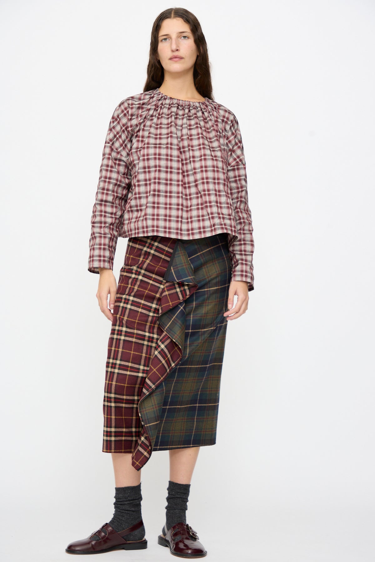 A person wears the Volant Skirt in Bordeaux and Military Green Check, a burgundy and green plaid wool midi skirt, with a ruffled front. Worn with a plaid top and burgundy loafers.