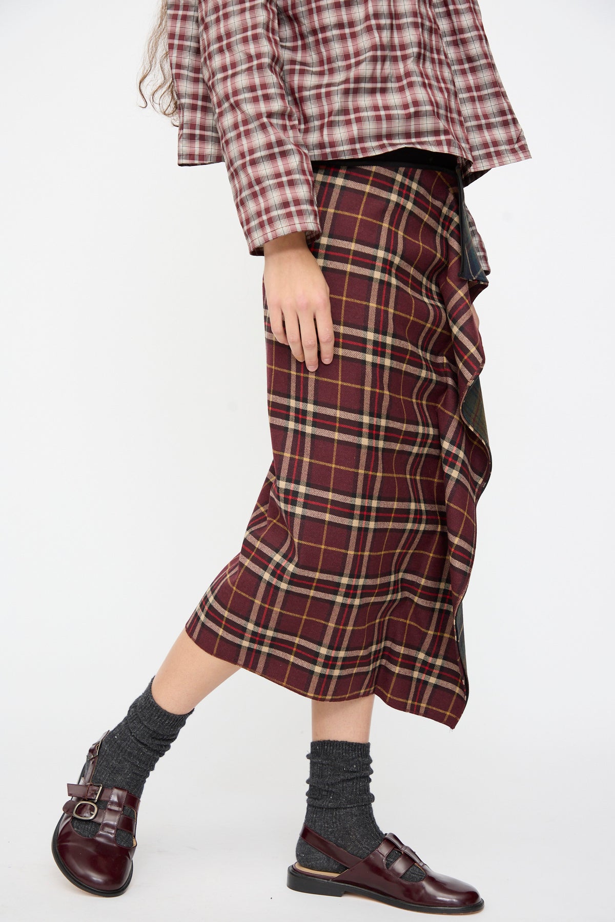 A person wearing an AVN Volant Skirt in Bordeaux and Military Green Check with a ruffled front detail, paired with a matching plaid shirt, grey socks, and dark red buckled shoes.