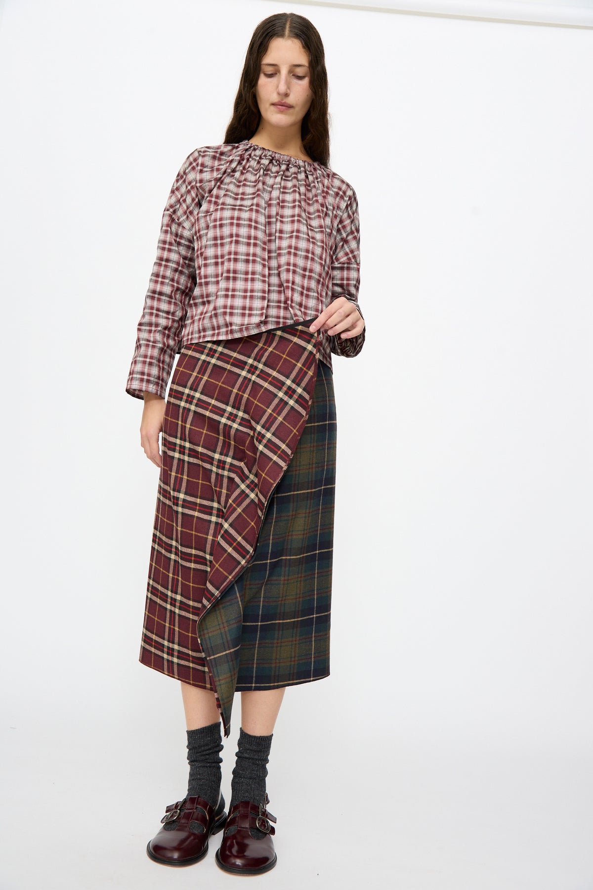 A person wears the Volant Skirt in Bordeaux and Military Green Check, a burgundy and green plaid wool midi skirt, with a ruffled front. Worn with a plaid top and burgundy loafers.