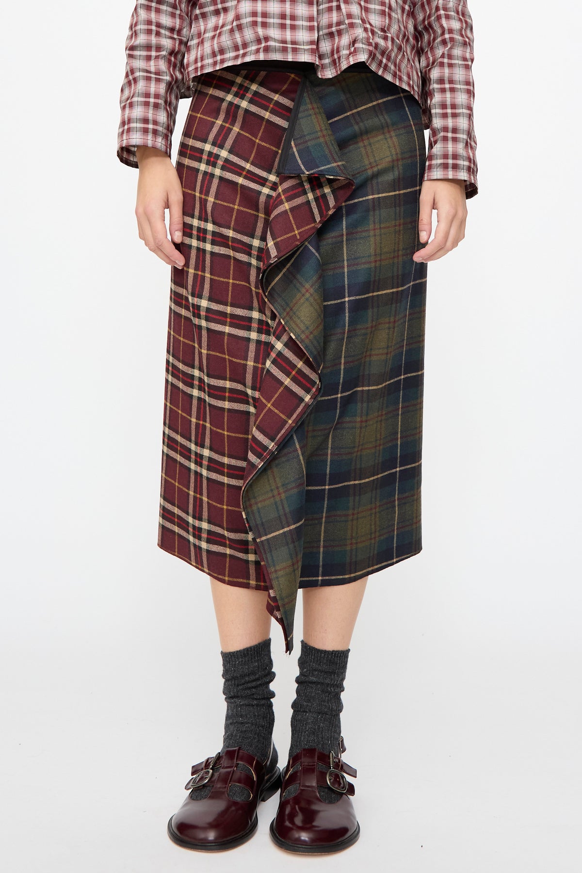 AVN Volant Skirt in Bordeaux and Military Green Check, a wool midi skirt with a ruffled vertical panel, worn with grey socks and dark buckled shoes.