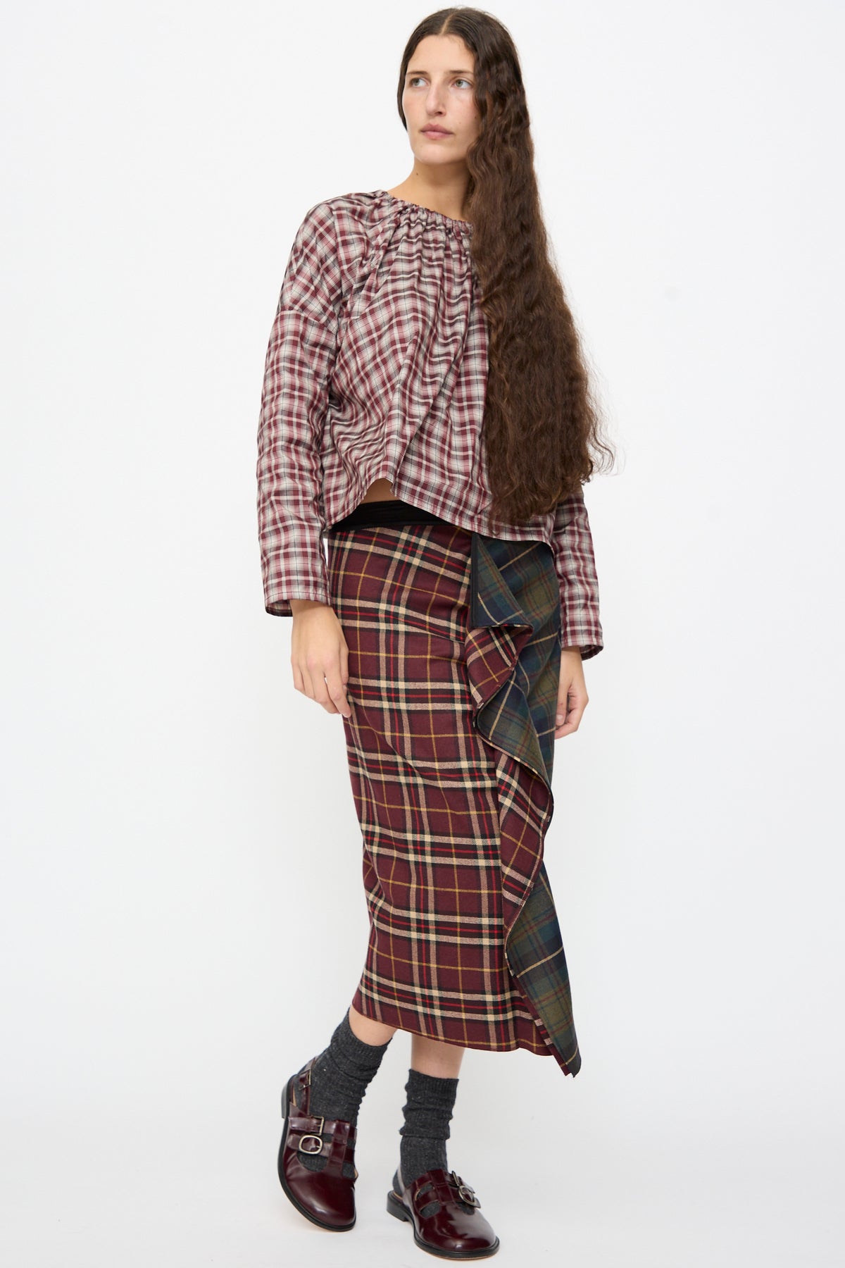 A person wears the Volant Skirt in Bordeaux and Military Green Check, a burgundy and green plaid wool midi skirt, with a ruffled front. Worn with a plaid top and burgundy loafers.