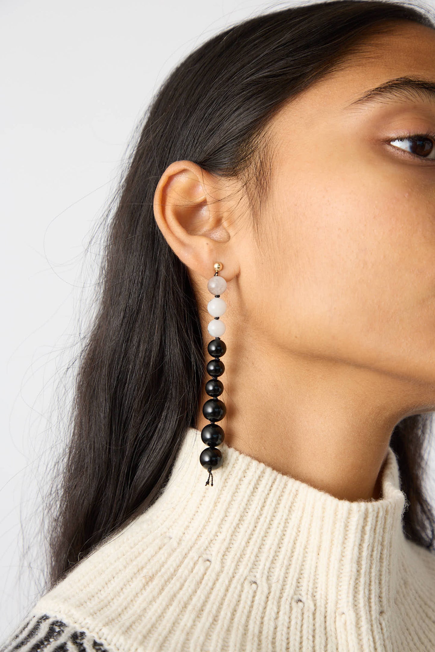 A close-up captures a person adorned in Abby Carnevale’s exquisite Gold Filled Beaded Earrings, featuring White Eagles Eye and Black Onyx beads. They have long dark hair and are wearing a light-colored sweater. These handmade earrings are crafted in NYC.