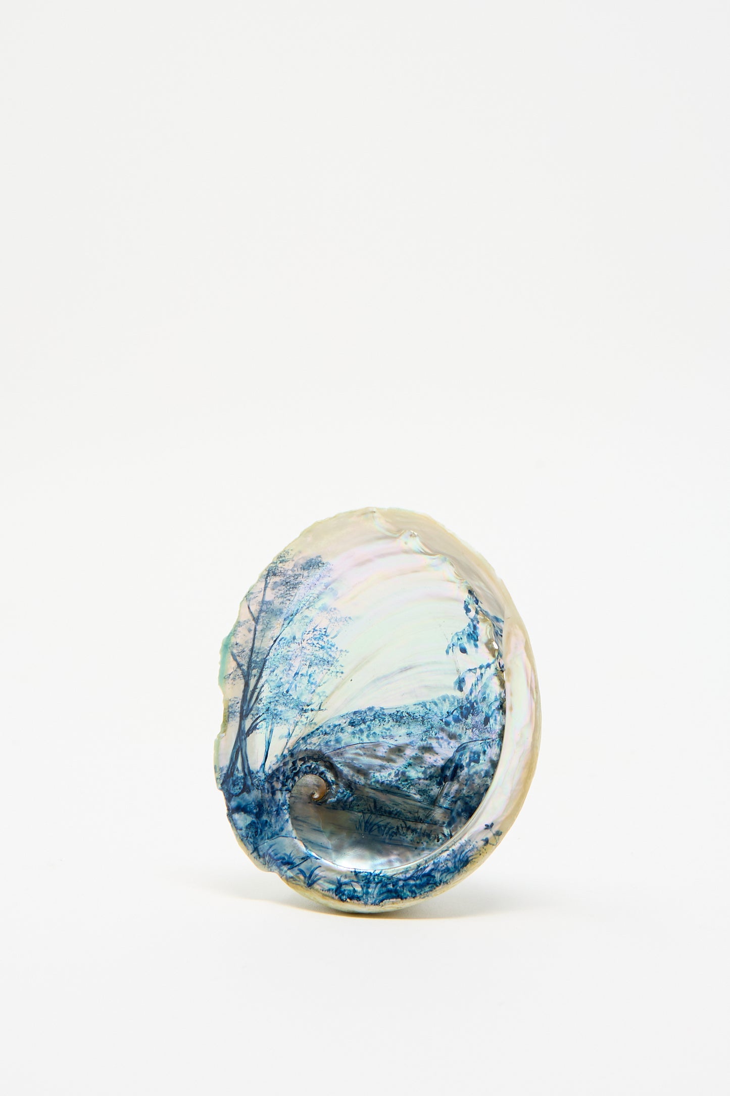 The "Hand Painted Abalone I" by Alyssa Goodman features a refined seashell, meticulously hand-painted with an intricate landscape of trees and hills, displayed inside a Abalone shell against a simple white background.