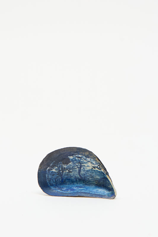 The "Hand Painted Steps Shell" by Alyssa Goodman features a dark blue seashell that evokes the appearance of abalone, with an intricate landscape painting set against a plain white background.