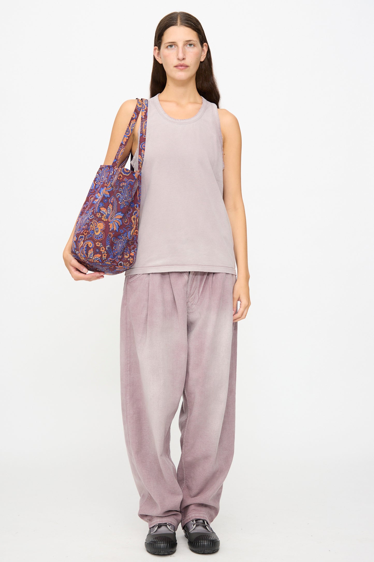 A woman stands facing forward wearing a light mauve tank top, loose mauve pants, black shoes, and carries the Amiacalva Easy Bag Medium in Paisley Azuki—a slouchy tote bag featuring a purple paisley-patterned flower print nylon, made in Japan.