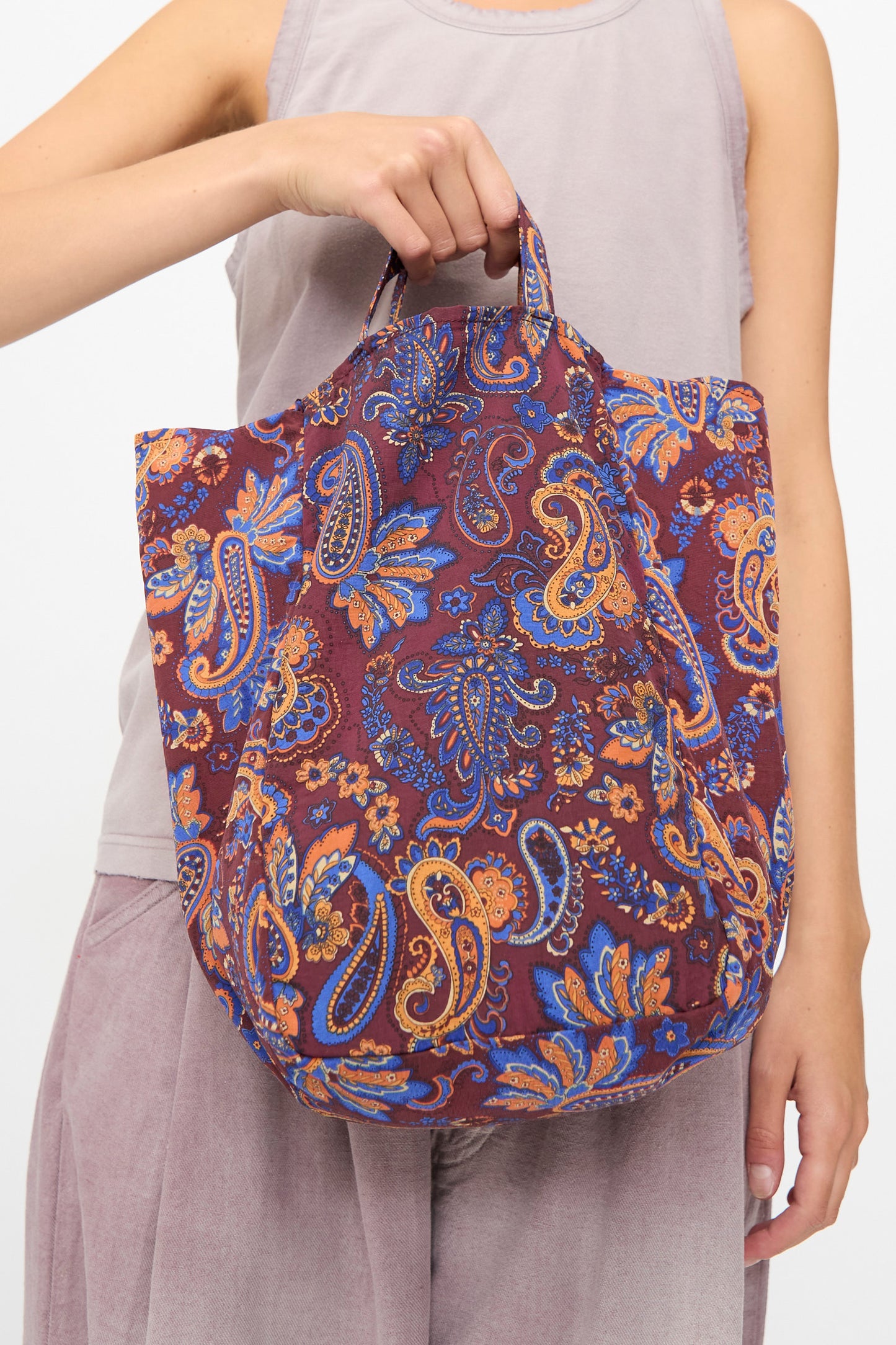 A person holding the Amiacalva Easy Bag Medium in Paisley Azuki—a slouchy tote bag featuring a paisley-patterned design in shades of purple, orange, and blue, made in Japan—paired with a sleeveless light mauve top and matching pants.