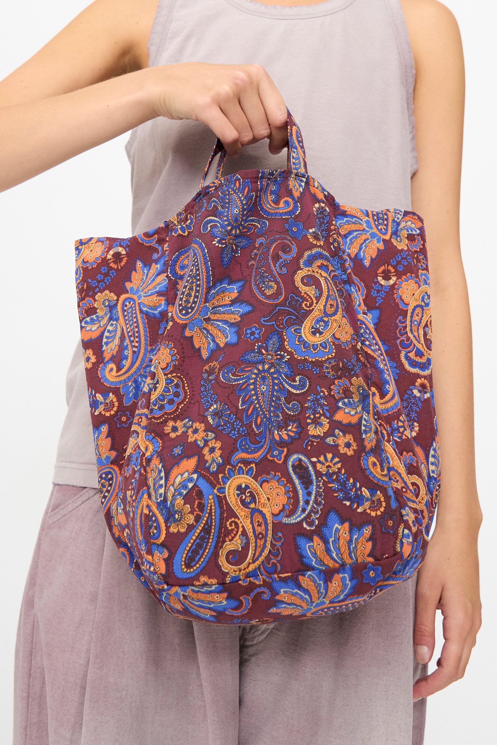 A person holding the Amiacalva Easy Bag Medium in Paisley Azuki—a slouchy tote bag featuring a paisley-patterned design in shades of purple, orange, and blue, made in Japan—paired with a sleeveless light mauve top and matching pants.