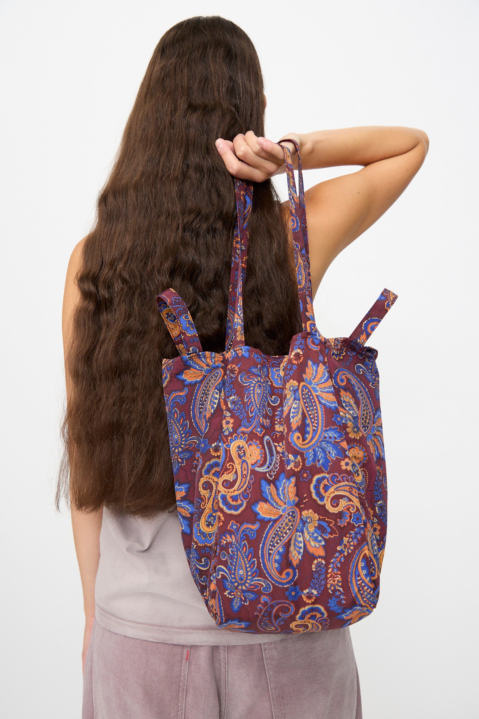 A person with long brown hair, seen from behind against a plain background, carries the Amiacalva Easy Bag Medium in Paisley Azuki slouchy tote over their shoulder. Crafted in Japan, this distinctive accessory blends quality craftsmanship with effortlessly stylish design.