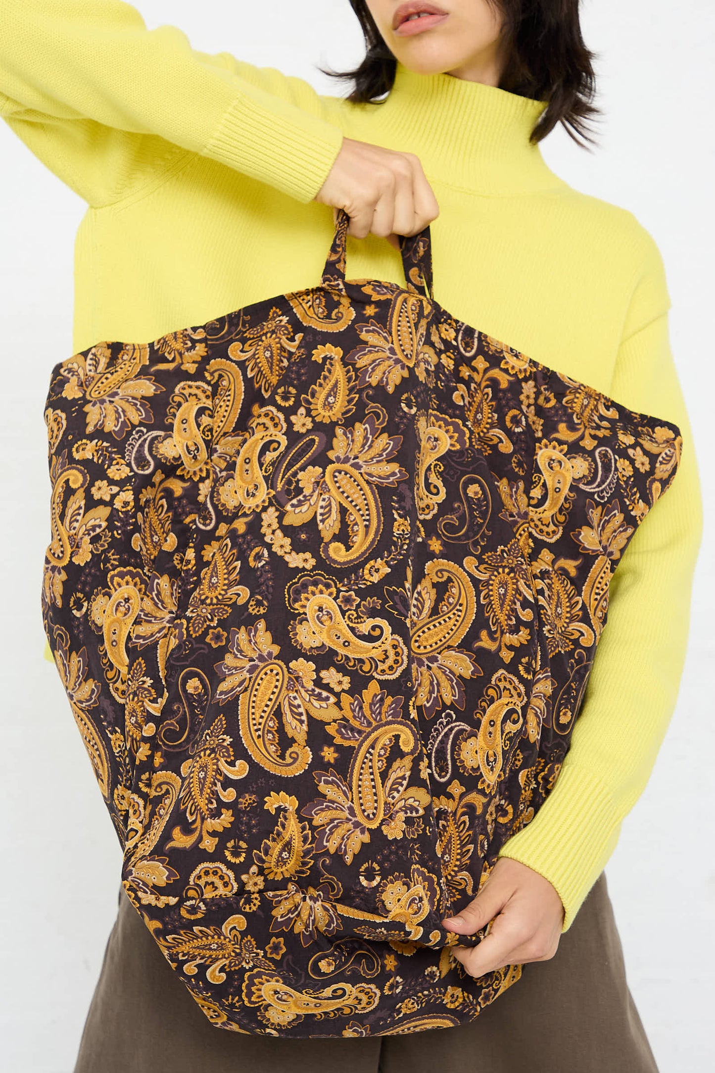 A person holding the Amiacalva Easy Large Bag in Paisley Brown pattern. The individual is wearing a yellow turtleneck sweater and gray pants.