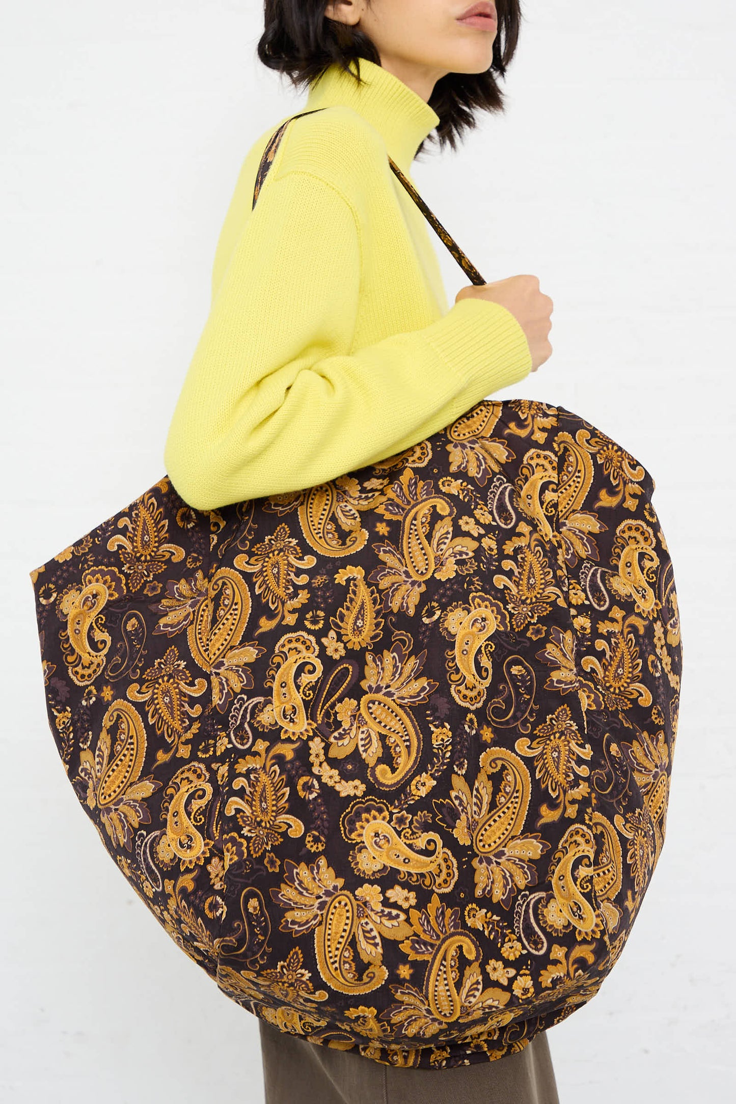 A person in a yellow sweater carries the Amiacalva Easy Large Bag in Paisley Brown.