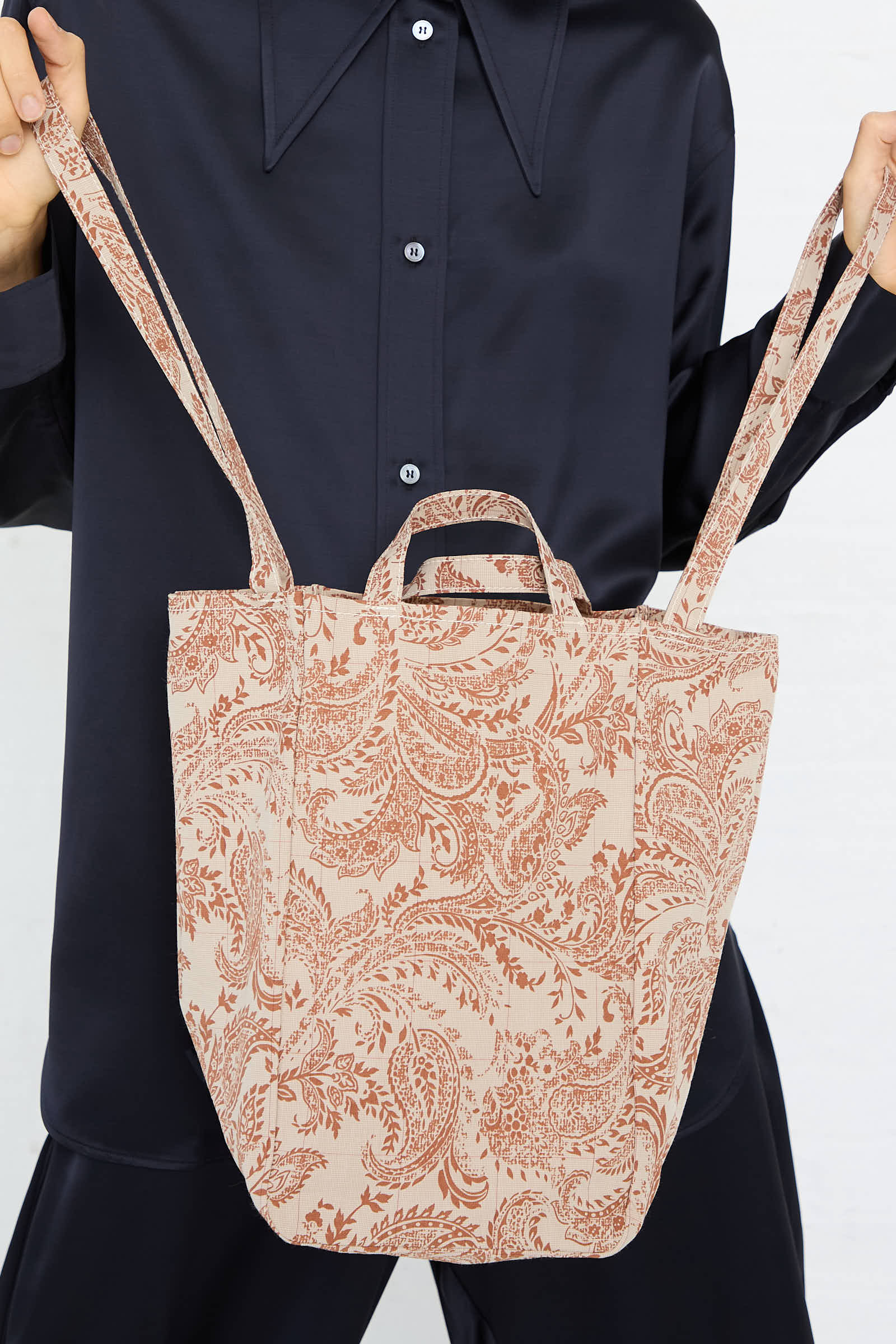 A person holds the Easy Medium Bag in Paisley Red by Amiacalva. Made in Japan, this beige slouchy tote bag features a red paisley pattern, two shoulder straps, and two small handles. The person wears a dark shirt.