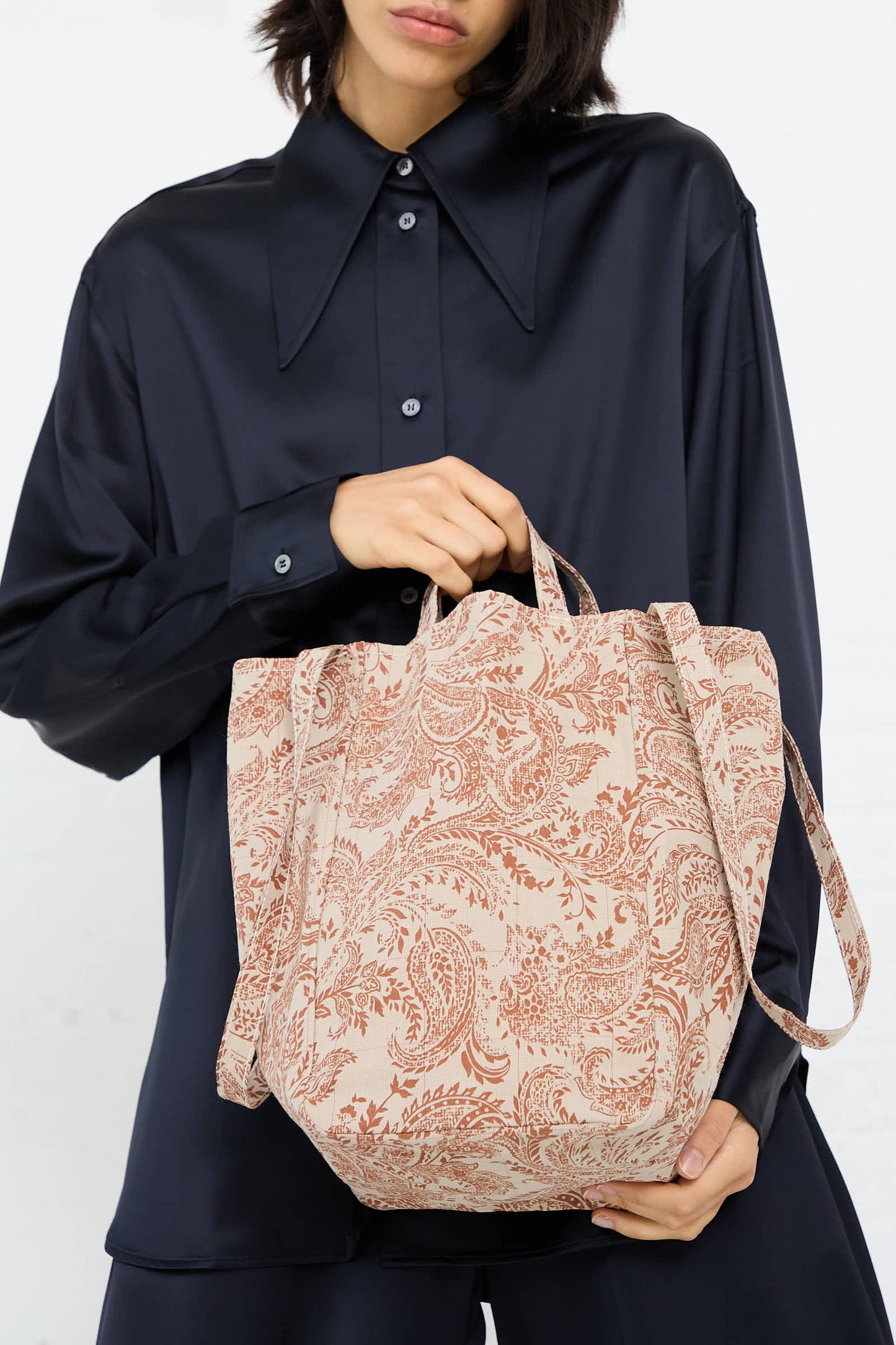 A person wearing a dark silky shirt holds an Amiacalva Easy Medium Bag in Paisley Red.