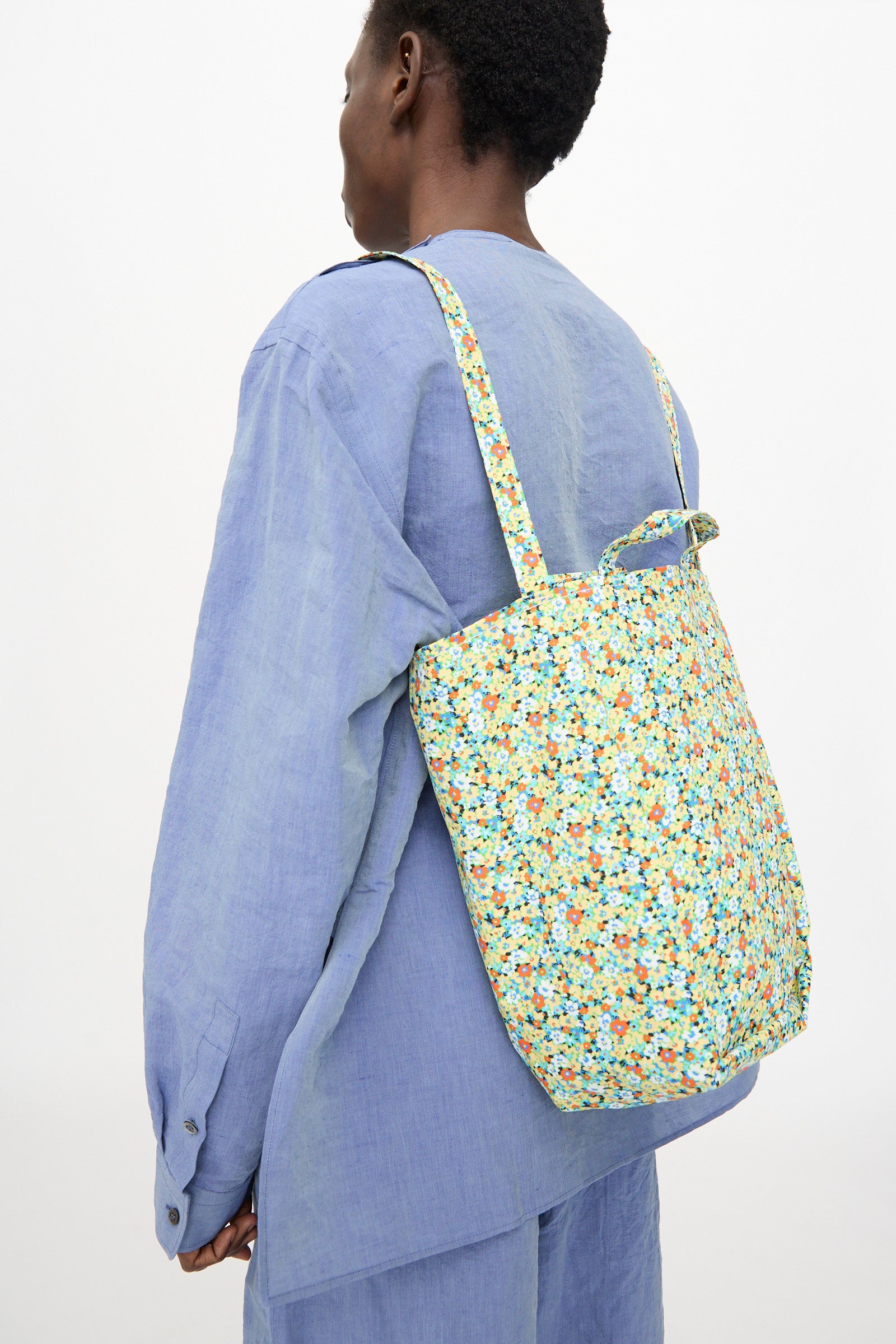 A person in a loose-fitting, light blue outfit carries the Amiacalva Medium Easy Bag in Flower Green— a flower-printed nylon slouchy tote made in Japan—over one shoulder, facing away from the camera against a plain background.