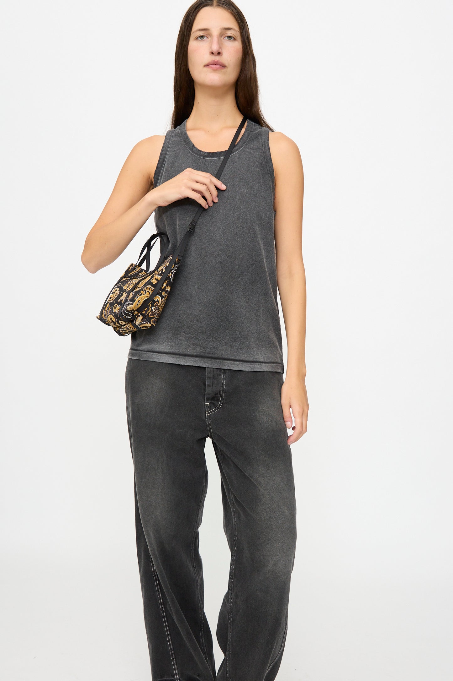 A woman stands against a plain white background, dressed in a sleeveless dark gray top and loose black jeans, while carrying the Amiacalva Two Way Shoulder Portable Bag in Paisley Black.