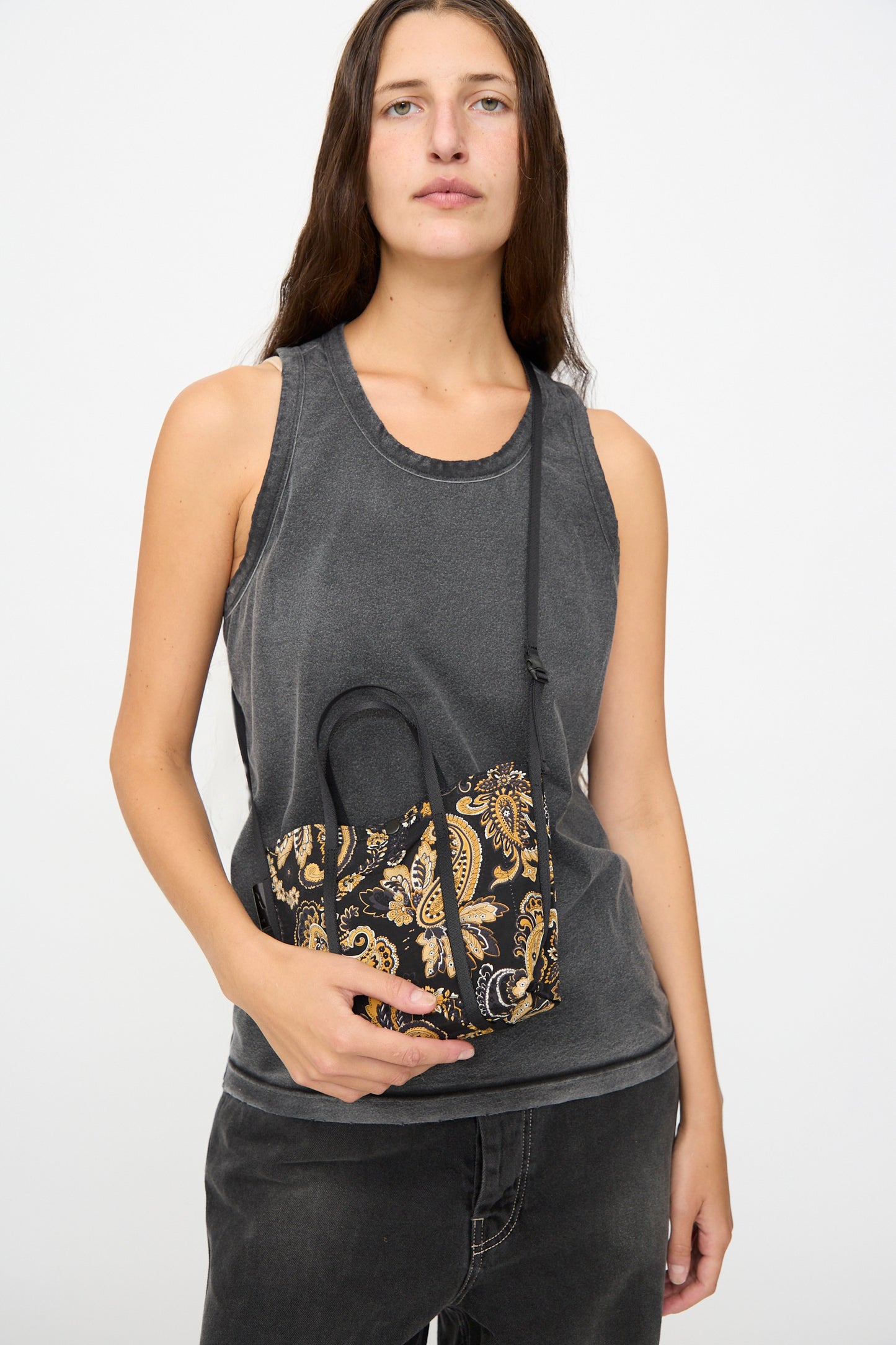 A woman in a sleeveless gray top and black jeans holds the Amiacalva Two Way Shoulder Portable Bag in Paisley Black, standing against a plain white background. Expertly crafted in Japan from nylon, this chic bag features gold accents and a striking paisley pattern.
