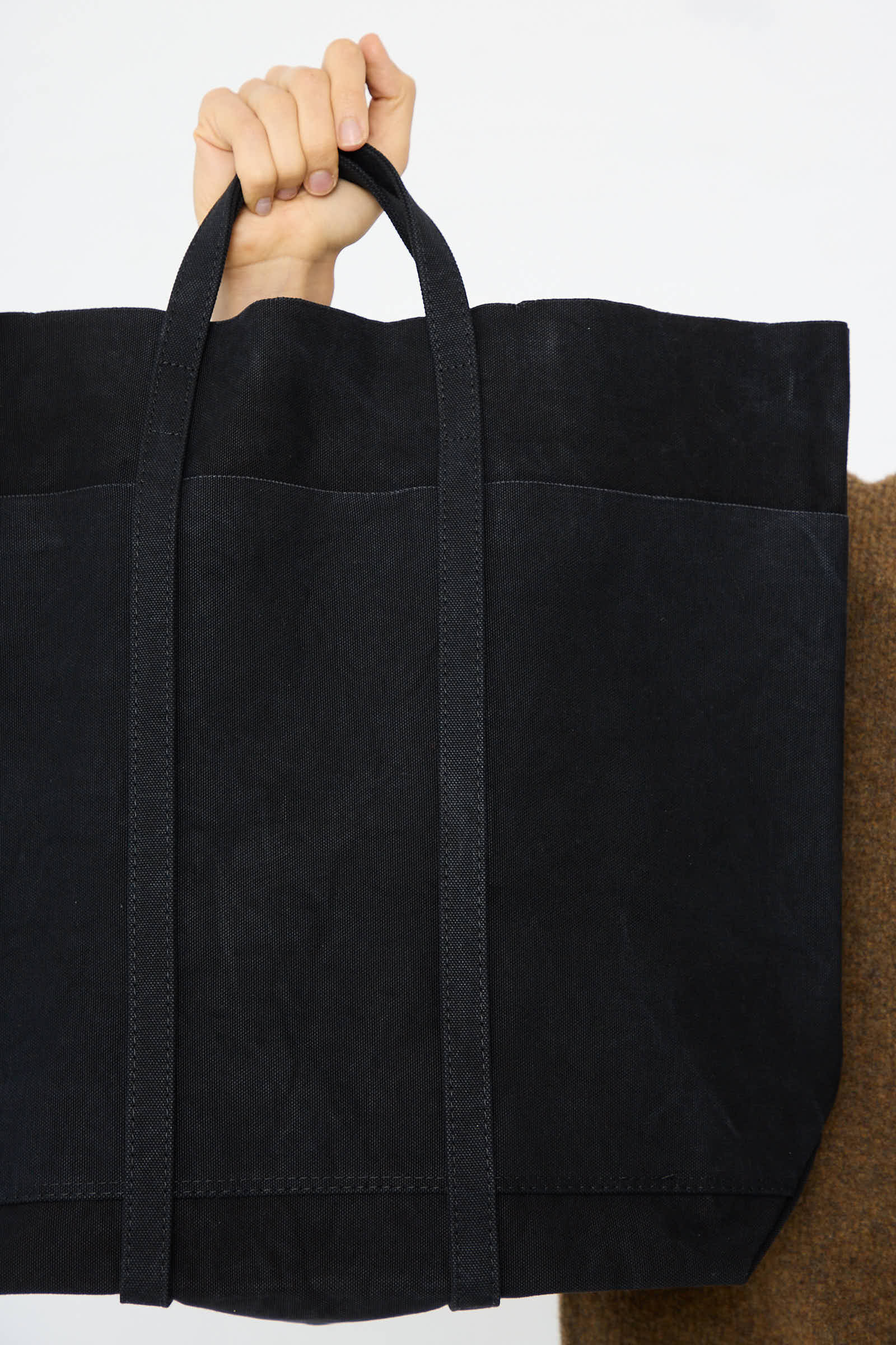 A hand holding the handles of a Washed Canvas 6 Pocket Tall Tote in Black by Amiacalva against a white background.