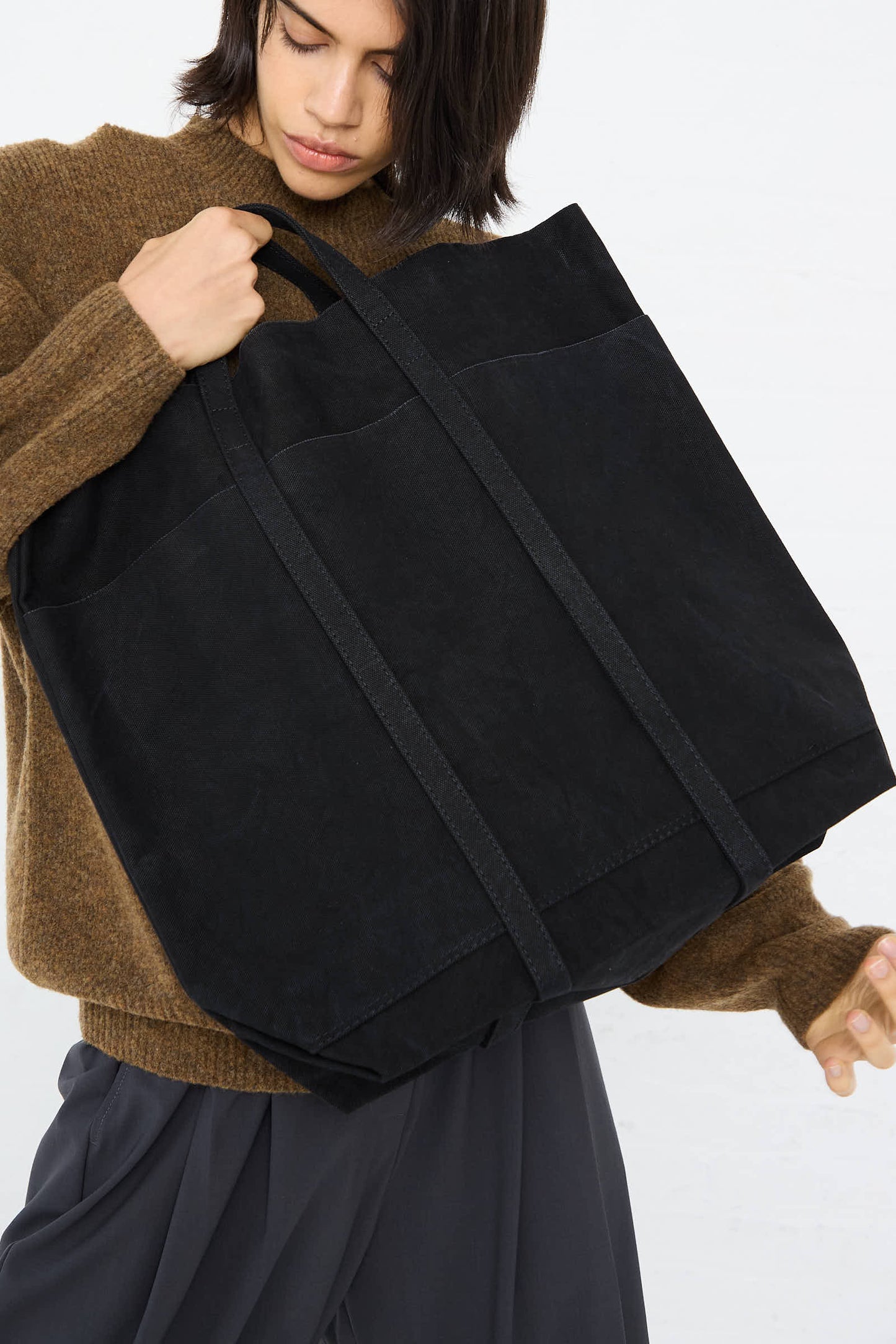 Person wearing a brown sweater and gray pants, holding an Amiacalva Washed Canvas 6 Pocket Tall Tote in Black.