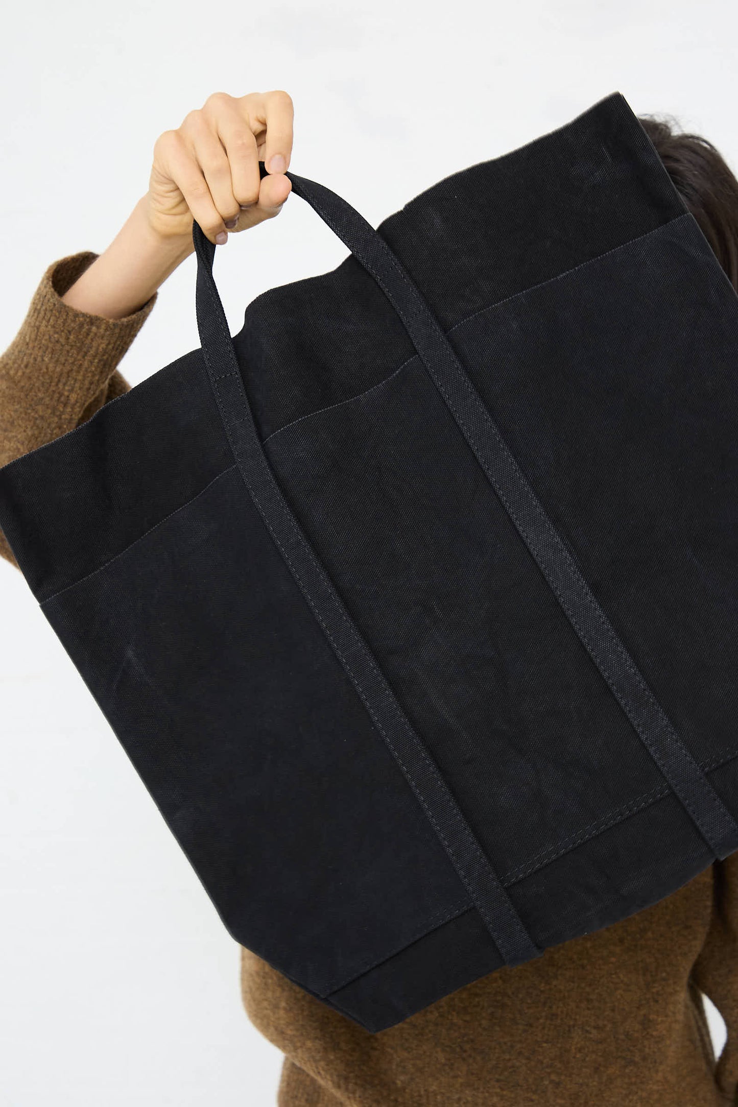 A person in a brown sweater holding up an Amiacalva Washed Canvas 6 Pocket Tall Tote in Black, partially covering their face.