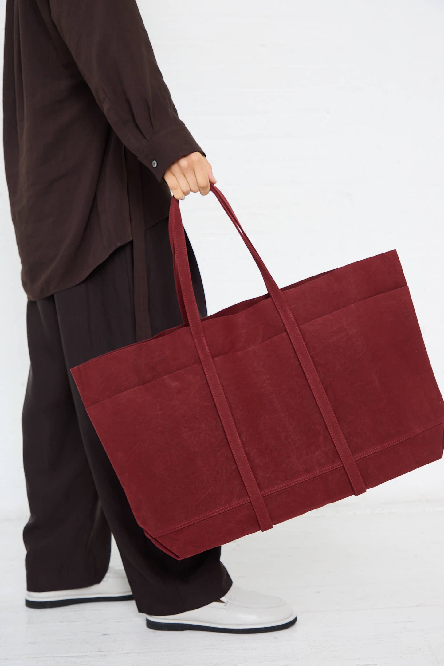 A person carrying the Amiacalva Washed Canvas 6 Pocket Large Tote in Burgundy with long handles, wearing dark clothing and white shoes against a white background.