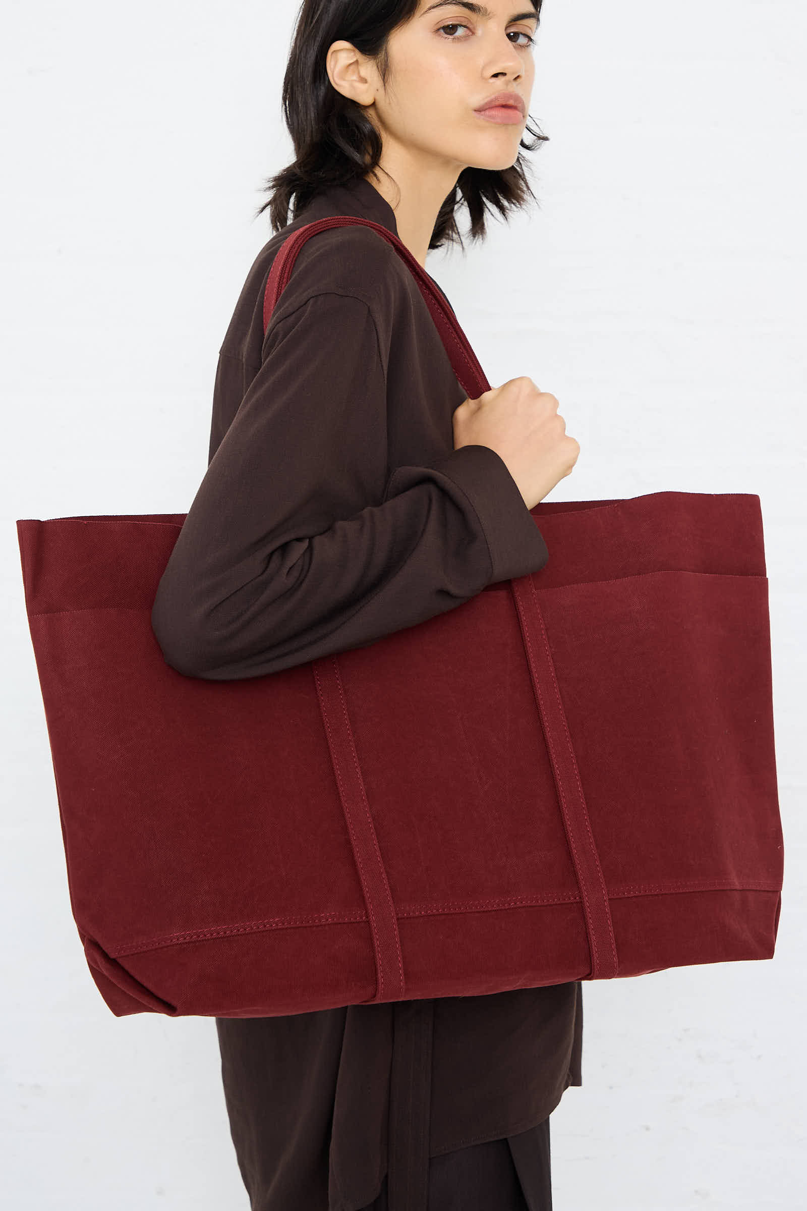 A person wearing a dark outfit, holding an Amiacalva Washed Canvas 6 Pocket Large Tote in Burgundy over their shoulder against a plain background.