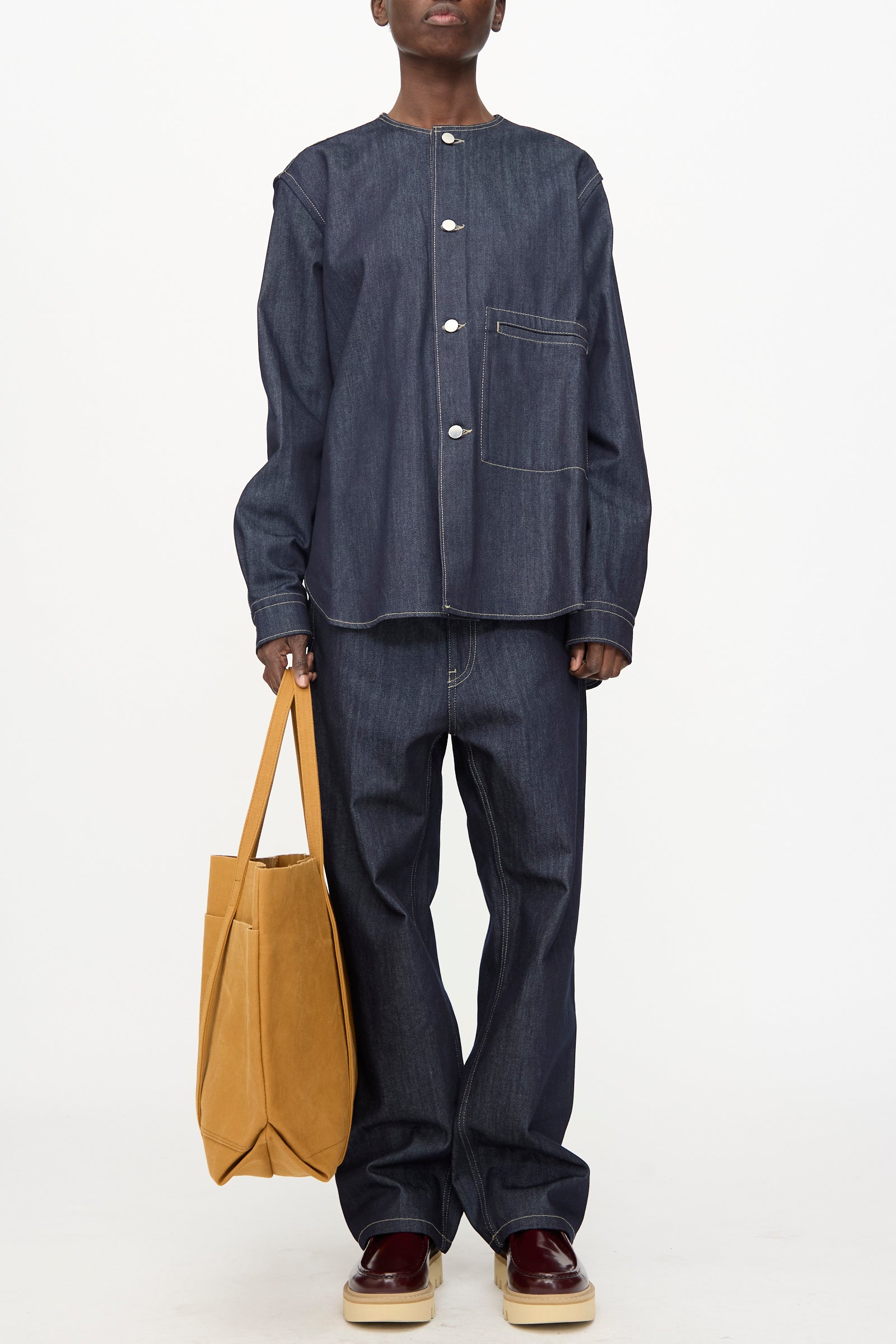Person wearing a loose dark denim shirt and jeans holds the Amiacalva Washed Canvas 6 Pockets Large Tote in Mustard made in Japan, standing in beige platform shoes with maroon accents against a plain background.