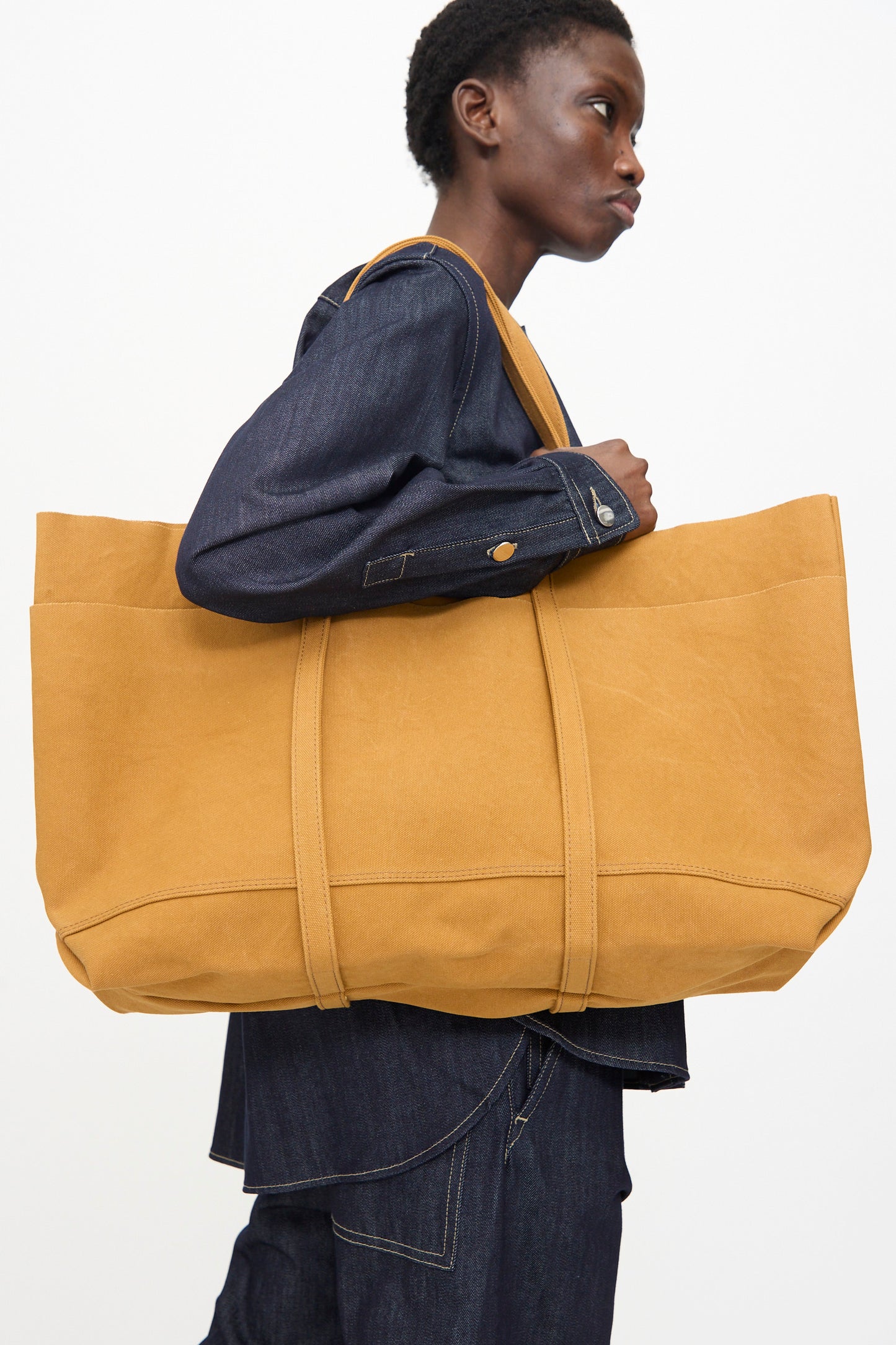 A person in dark denim carries the Amiacalva Washed Canvas 6 Pockets Large Tote in Mustard, made in Japan, over their shoulder against a plain background.