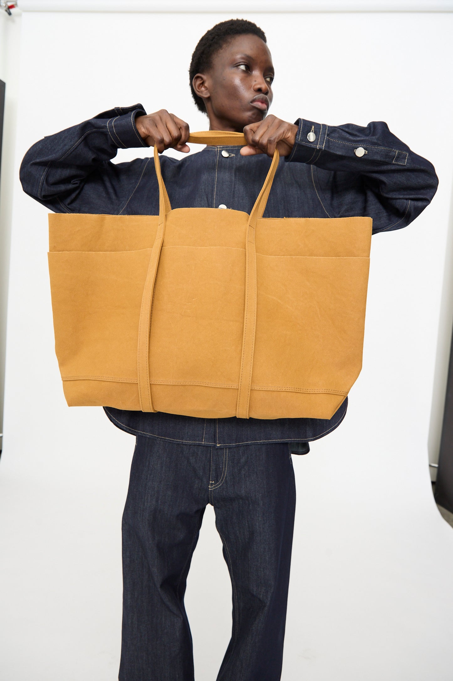 A person in dark denim holds the Amiacalva Washed Canvas 6 Pockets Large Tote in Mustard, made in Japan, against a plain white background.