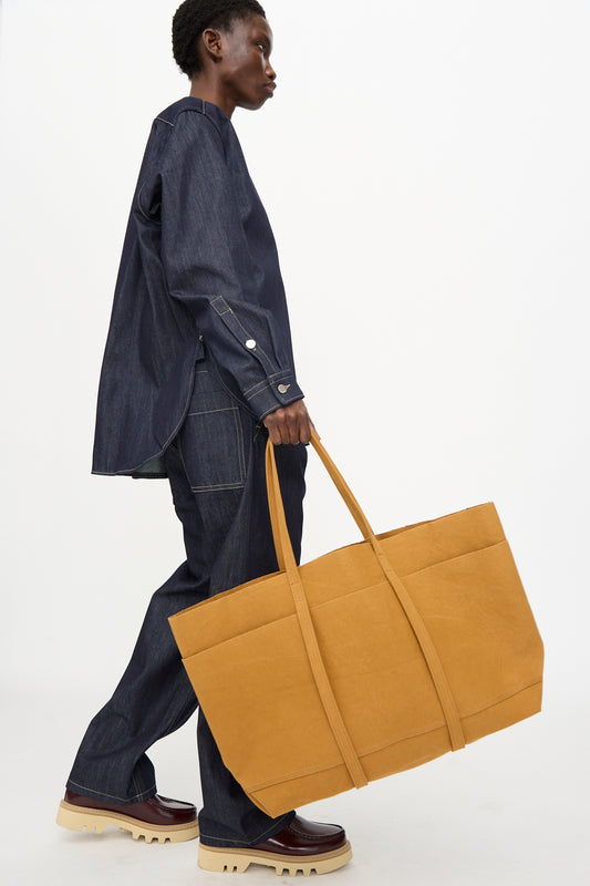 A person in dark denim and chunky shoes walks with the Amiacalva Washed Canvas 6 Pockets Large Tote in Mustard, its oversized Japanese-made design popping against the plain background.