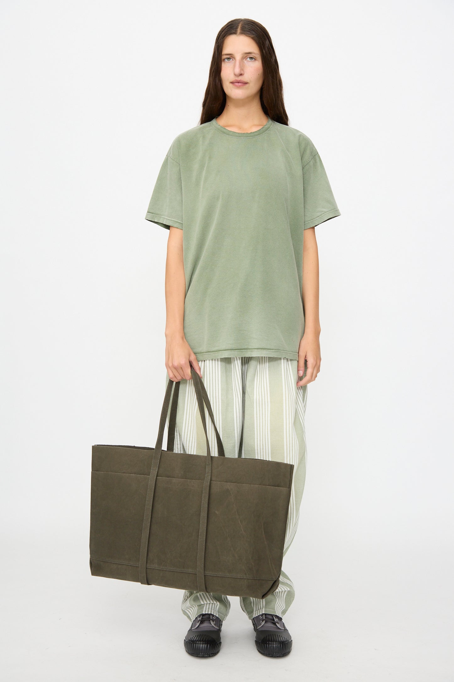 Against a white background, the individual wears a loose green t-shirt, striped pants, and black shoes, complemented by the Amiacalva Washed Canvas 6 Pockets Large Tote in Olive.