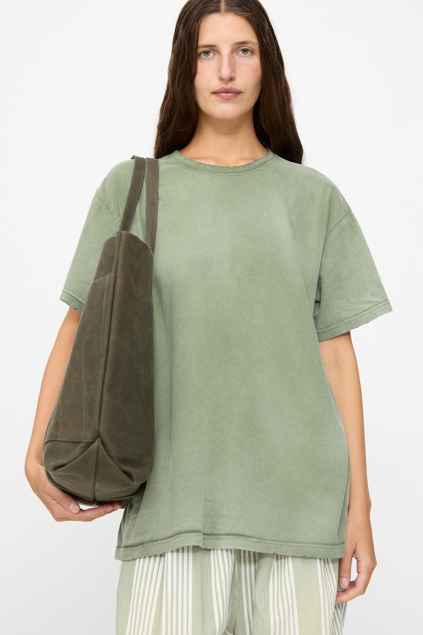 A person with long brown hair wears a loose green T-shirt and striped pants, carrying the Amiacalva Washed Canvas 6 Pockets Large Tote in Olive, standing against a plain white background.