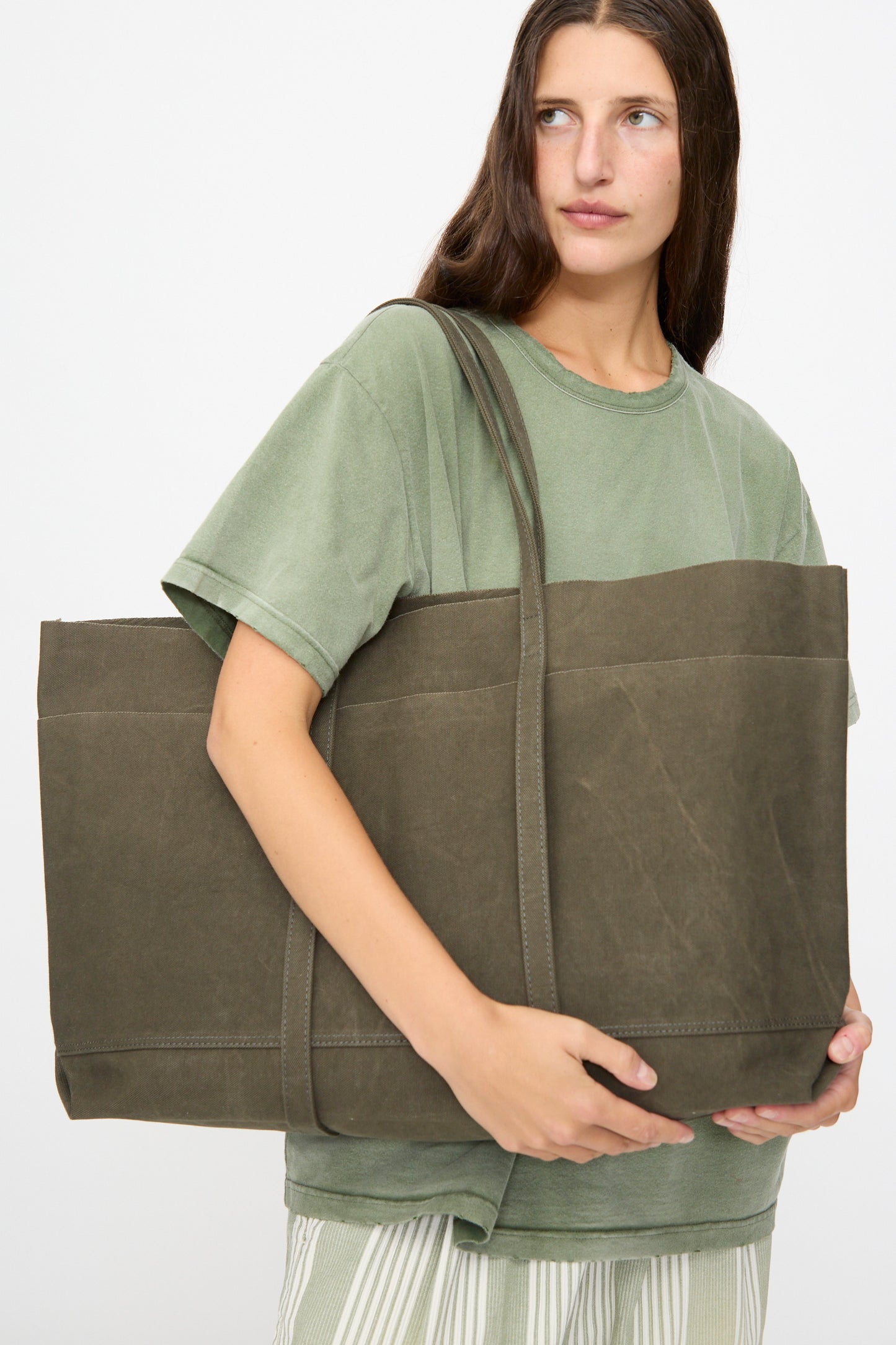 A person with long brown hair holds the Amiacalva Washed Canvas 6 Pockets Large Tote in Olive—crafted in Japan—while wearing a light green t-shirt and striped pants against a plain white background.