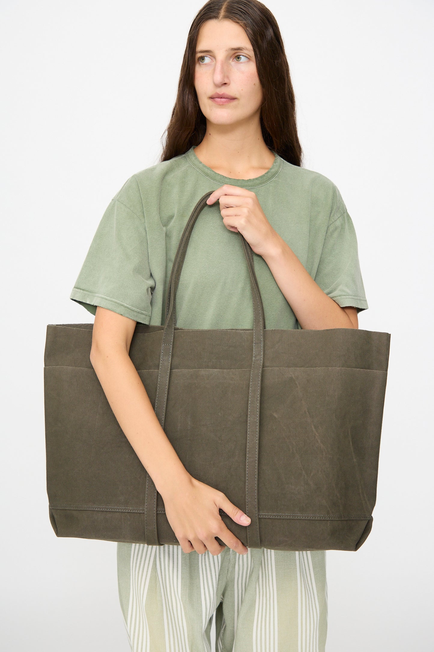 A woman wearing a green t-shirt and striped pants stands against a plain background, holding the Amiacalva Washed Canvas 6 Pockets Large Tote in Olive with both hands. Made in Japan, this tote blends classic design with practical functionality.