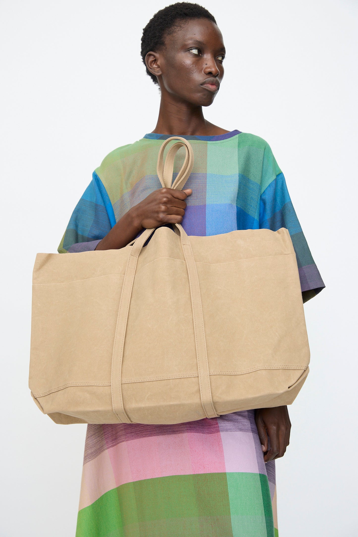 A person holds the Amiacalva Washed Canvas 6 Pockets Large Tote in Taupe against a plain white background, wearing a colorful plaid dress.