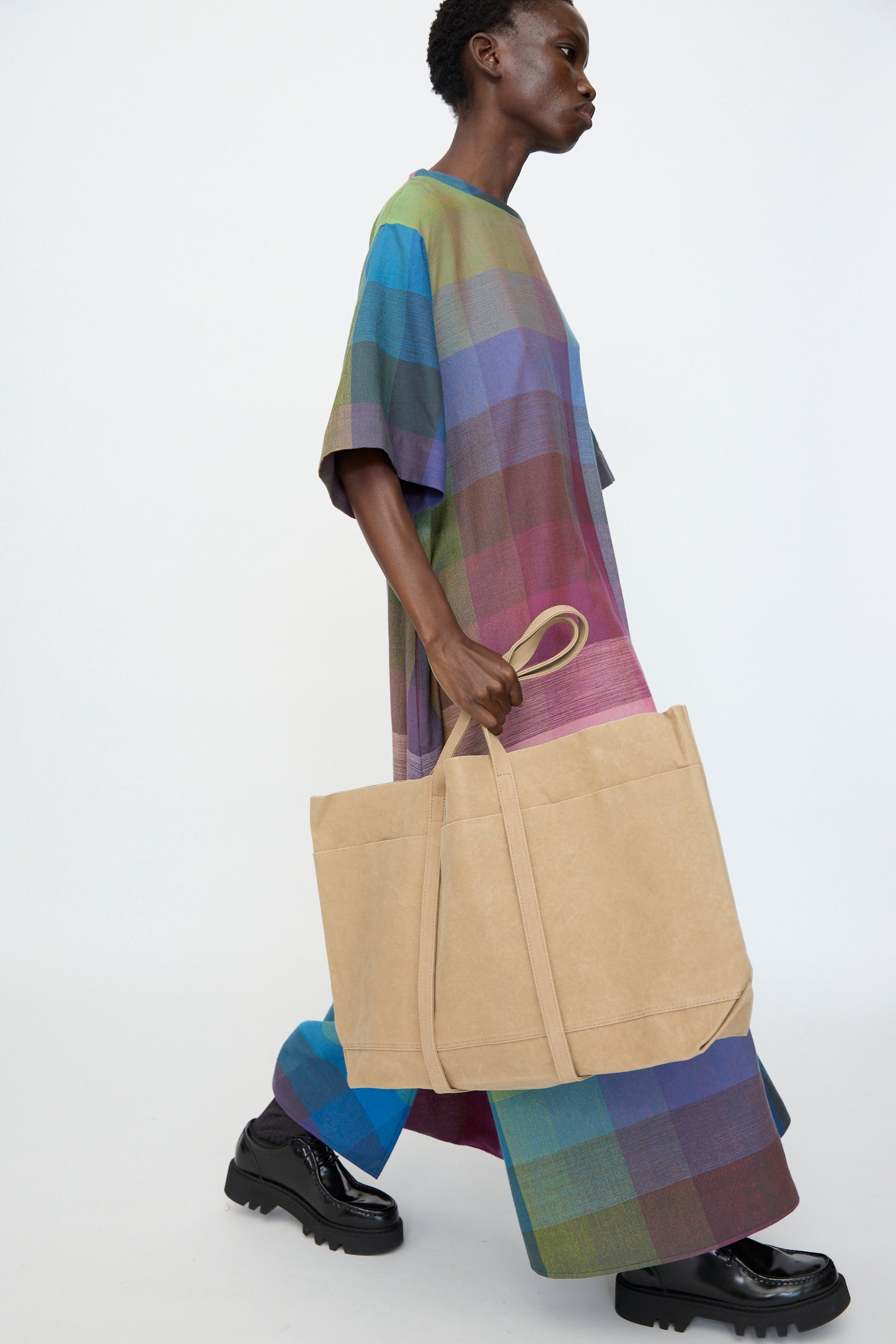 A person walks against a plain background, carrying the Amiacalva Washed Canvas 6 Pockets Large Tote in Taupe, wearing a loose multicolored plaid dress and black shoes.