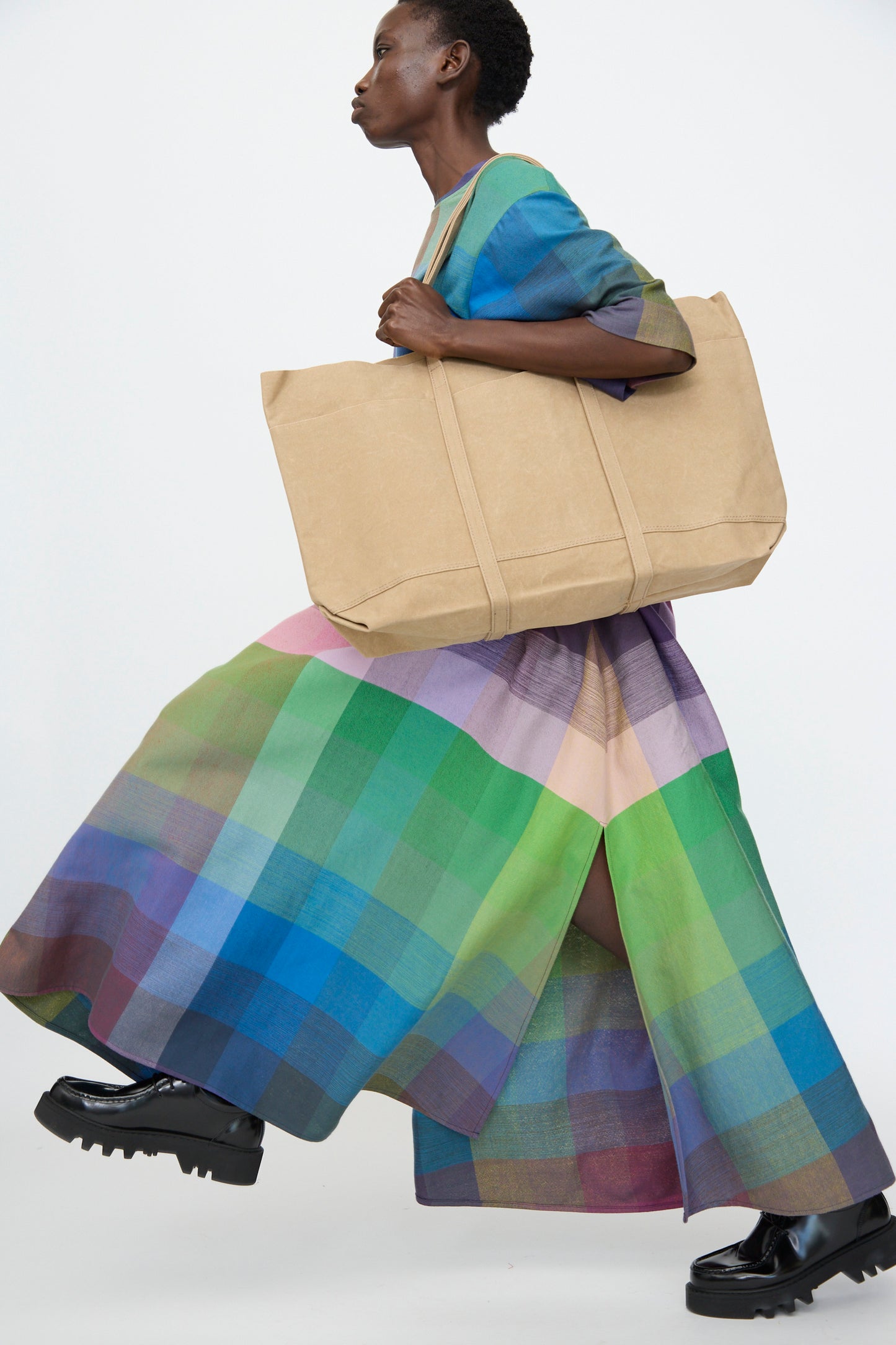 A person carries the Amiacalva Washed Canvas 6 Pockets Large Tote in Taupe, paired with a long colorful plaid dress and black shoes, walking against a plain background.