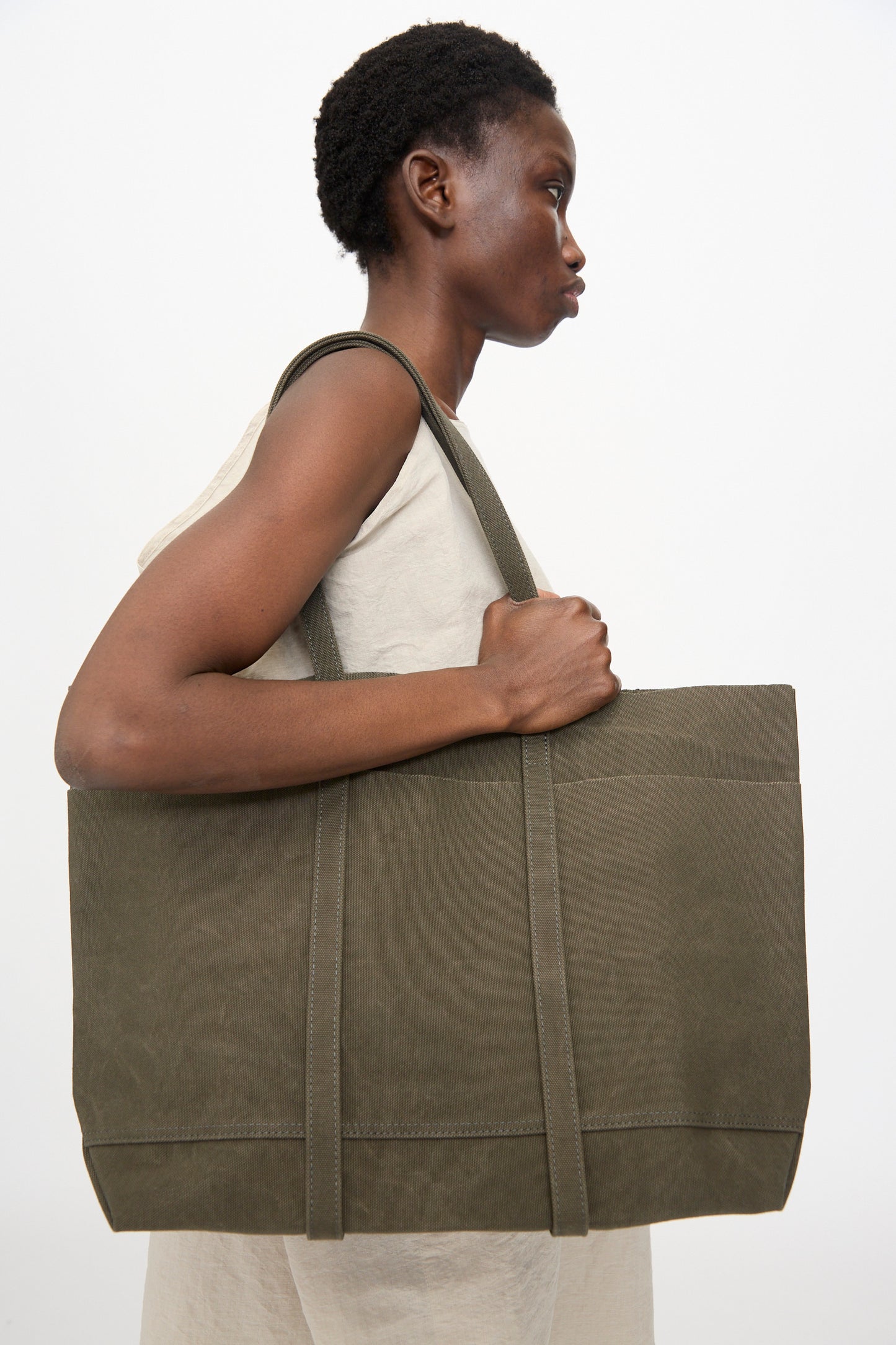 A person in a sleeveless beige top carries the Amiacalva Washed Canvas 6 Pockets Medium Tote in Olive, made in Japan, over their shoulder while standing against a plain white background.