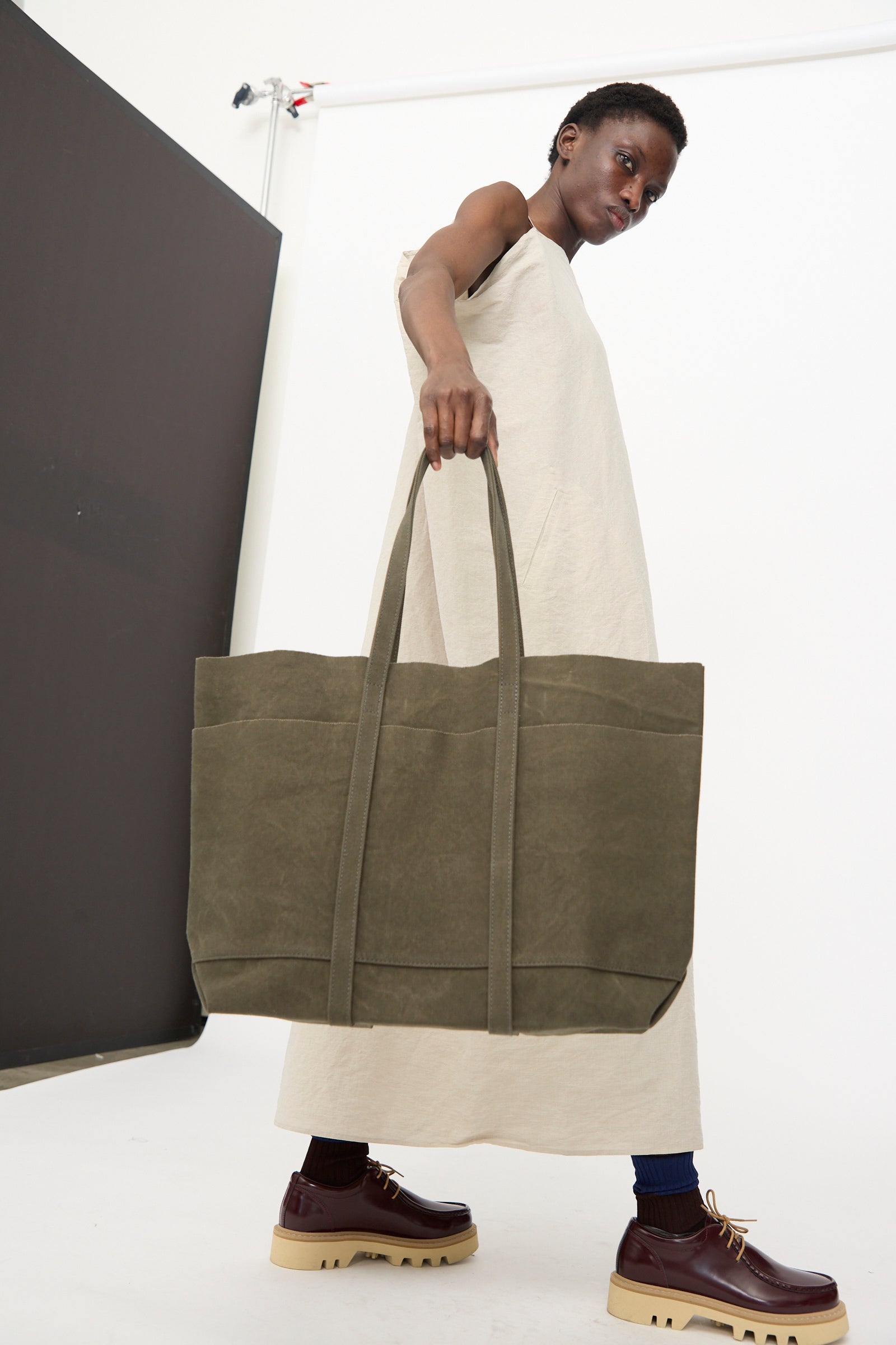 A person in a sleeveless beige dress holds the Amiacalva Washed Canvas 6 Pockets Medium Tote in Olive, made in Japan, and wears maroon platform shoes with tan soles against a minimal studio backdrop.