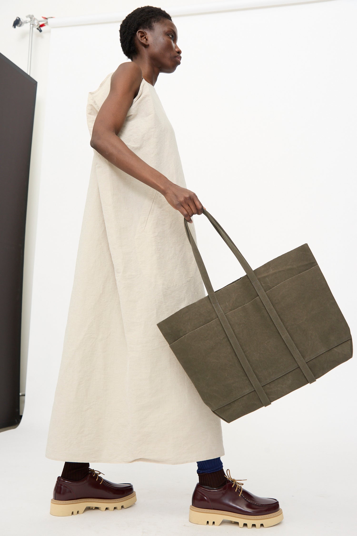 A person in a sleeveless, ankle-length beige dress holds the Amiacalva Washed Canvas 6 Pockets Medium Tote in Olive and wears burgundy platform shoes with beige soles.