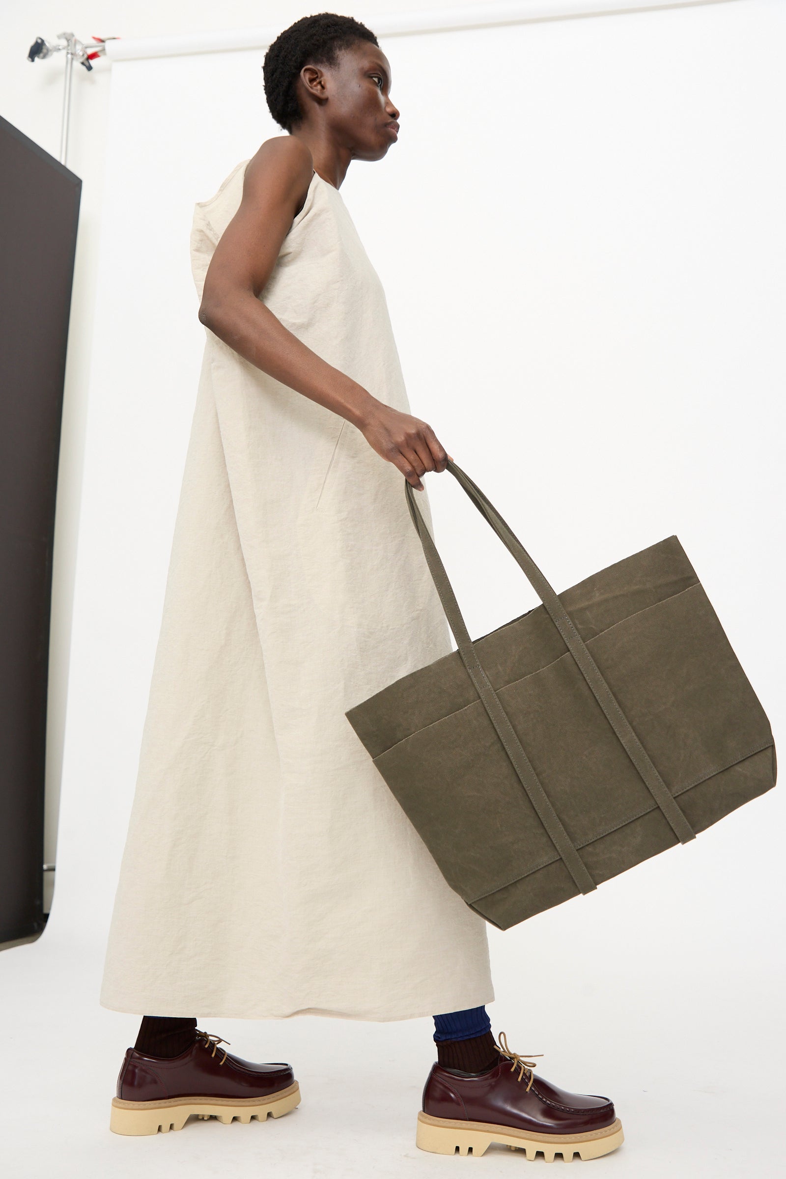A person in a sleeveless, ankle-length beige dress holds the Amiacalva Washed Canvas 6 Pockets Medium Tote in Olive and wears burgundy platform shoes with beige soles.