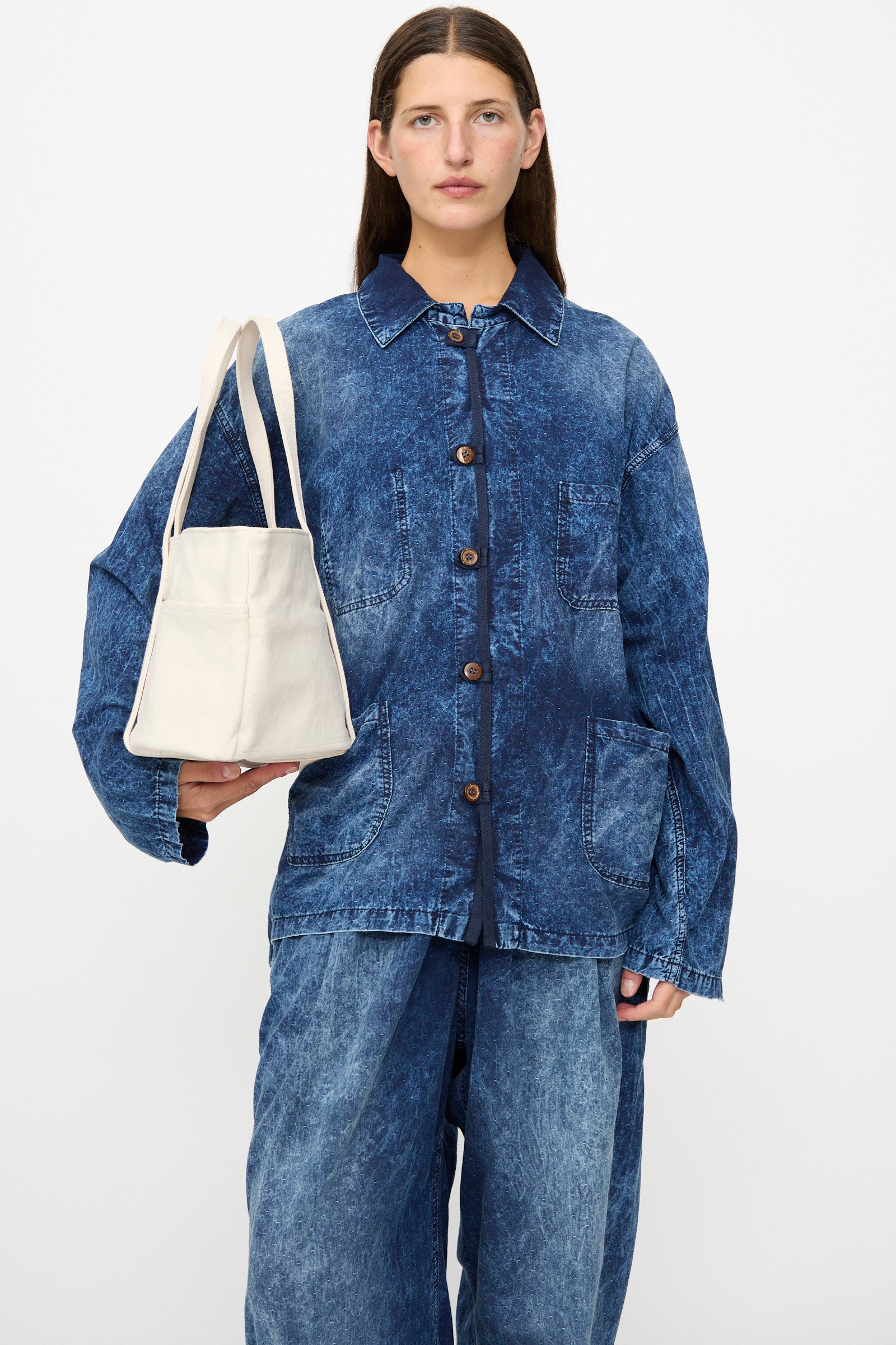 A person wearing a loose-fitting, button-up blue denim jacket and matching pants holds the Amiacalva Washed Canvas 6 Pockets Medium Tote in White, made in Japan, against a plain white background.