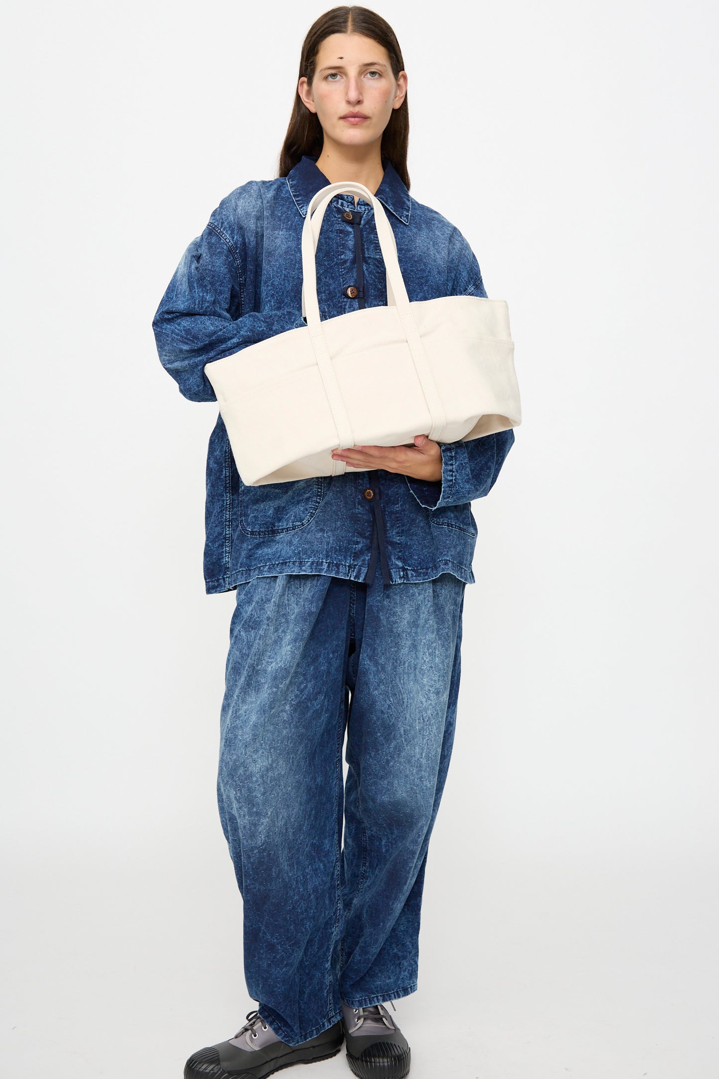 A person wearing a loose blue denim jacket and pants holds the Amiacalva Washed Canvas 6 Pockets Medium Tote in White against a plain white background.
