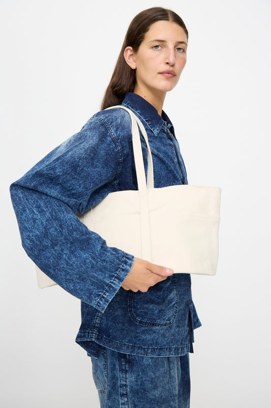 A person dressed in blue denim holds the Amiacalva Washed Canvas 6 Pockets Medium Tote in White—crafted in Japan from plain, cream-colored washed canvas—under their arm, looking forward against a plain light background.