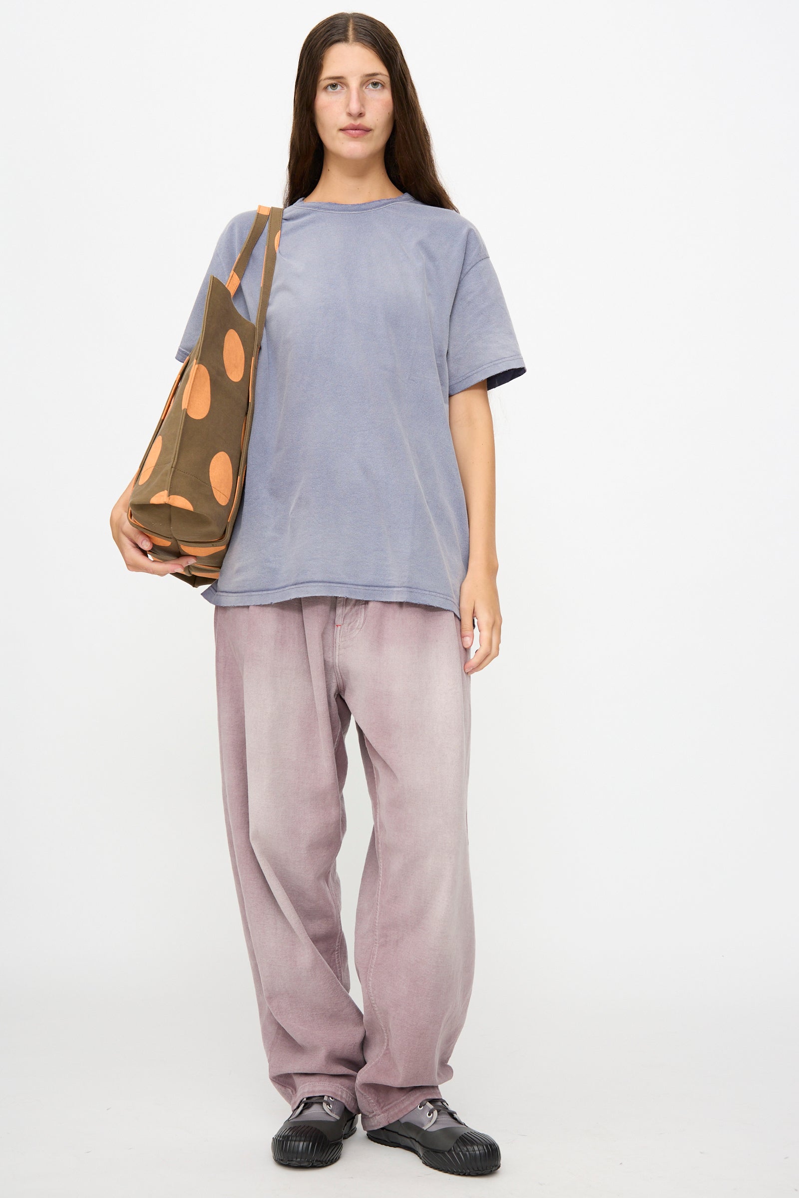 A person stands facing forward, wearing a loose light blue t-shirt, baggy light purple pants, black shoes, and carrying the Amiacalva Washed Canvas Medium Dot Tote in Orange.