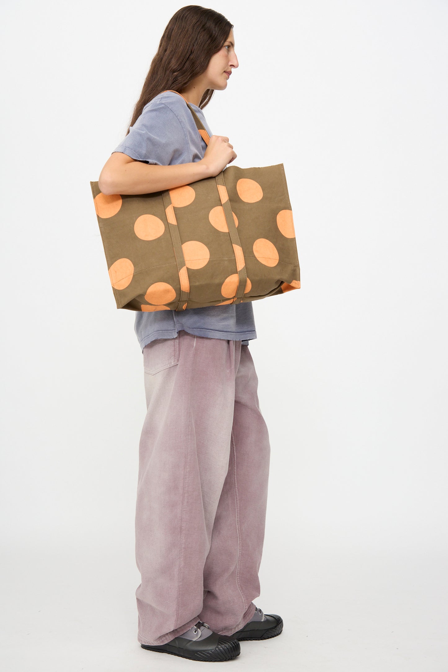 A person stands in profile against a plain white background, wearing a grey shirt and loose pink pants, holding the Amiacalva Washed Canvas Medium Dot Tote in Orange.