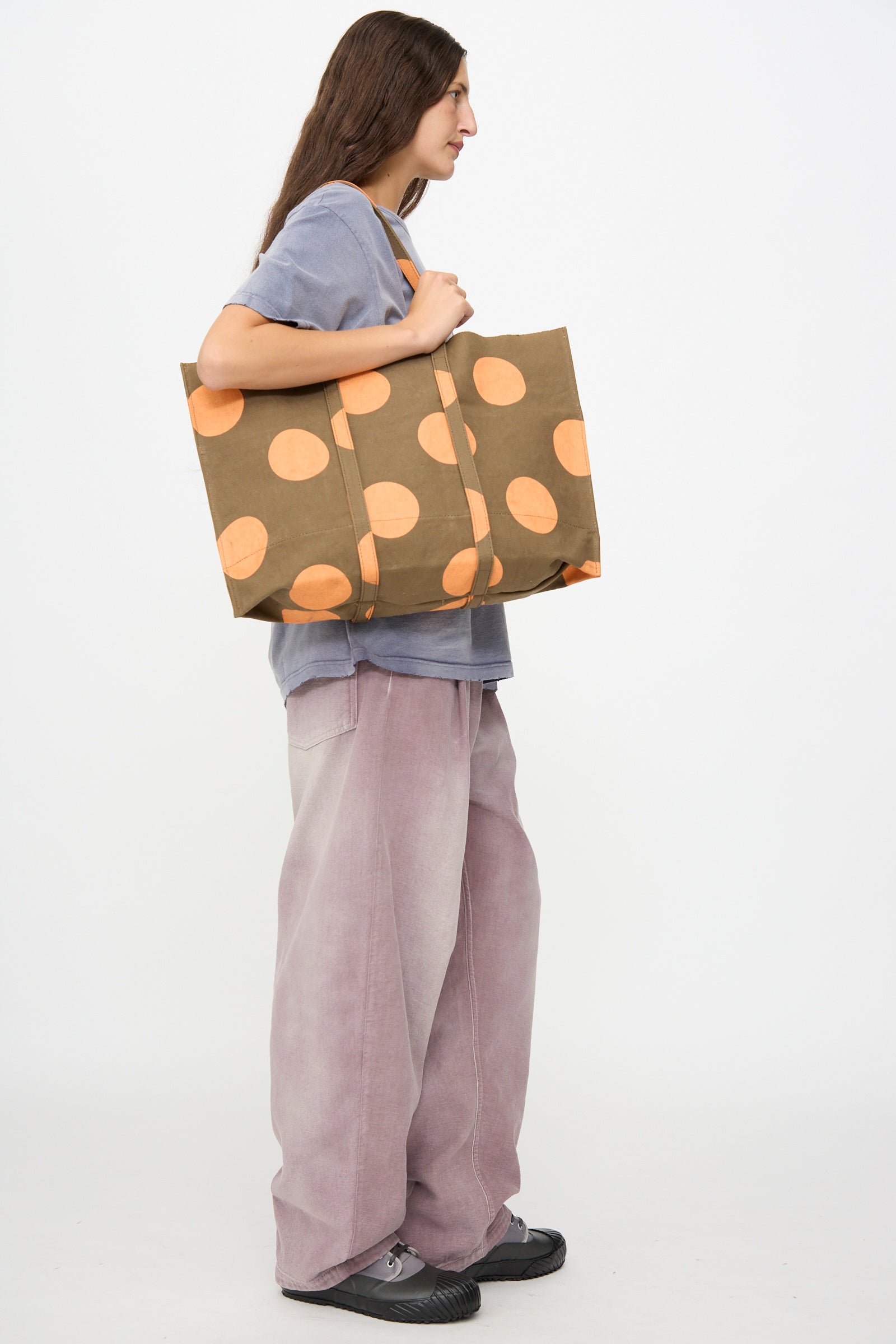 A person stands in profile against a plain white background, wearing a grey shirt and loose pink pants, holding the Amiacalva Washed Canvas Medium Dot Tote in Orange.