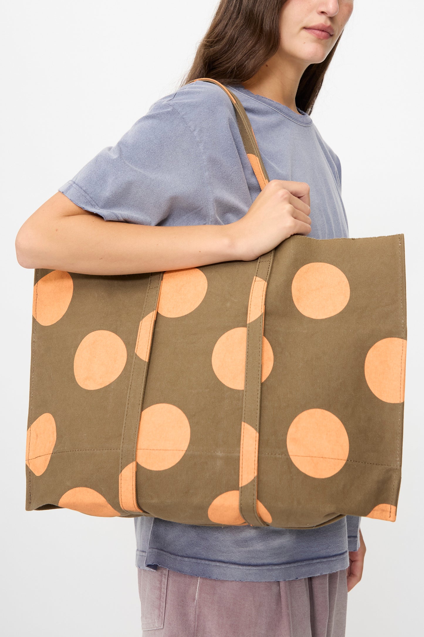 A person, shown from the shoulders down, carries the Amiacalva Washed Canvas Medium Dot Tote in Orange—an olive green bag with orange polka dots—while wearing a short-sleeve gray shirt and mauve pants.