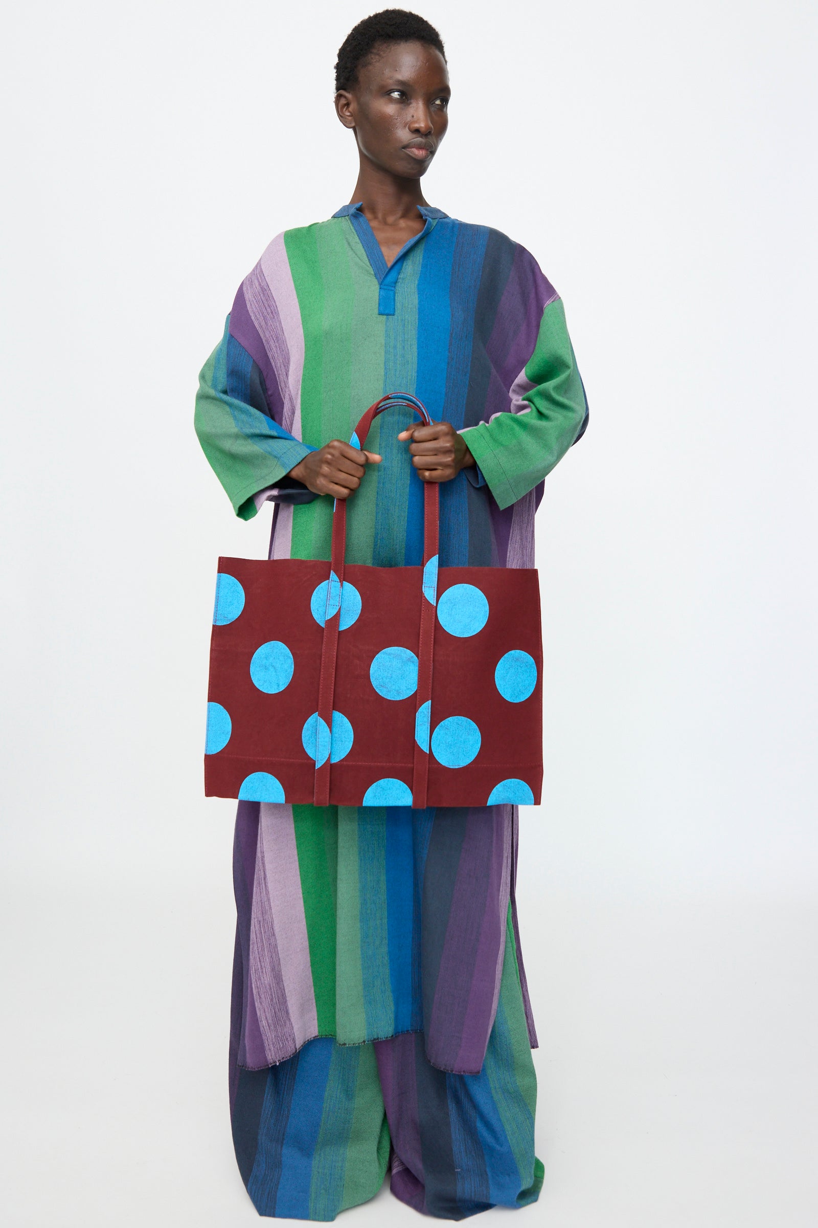 A person in a loose, striped, multi-colored outfit holds the Amiacalva Washed Canvas Medium Dot Tote in Sky Blue against a plain white background. This charming tote brings playful color to the scene.