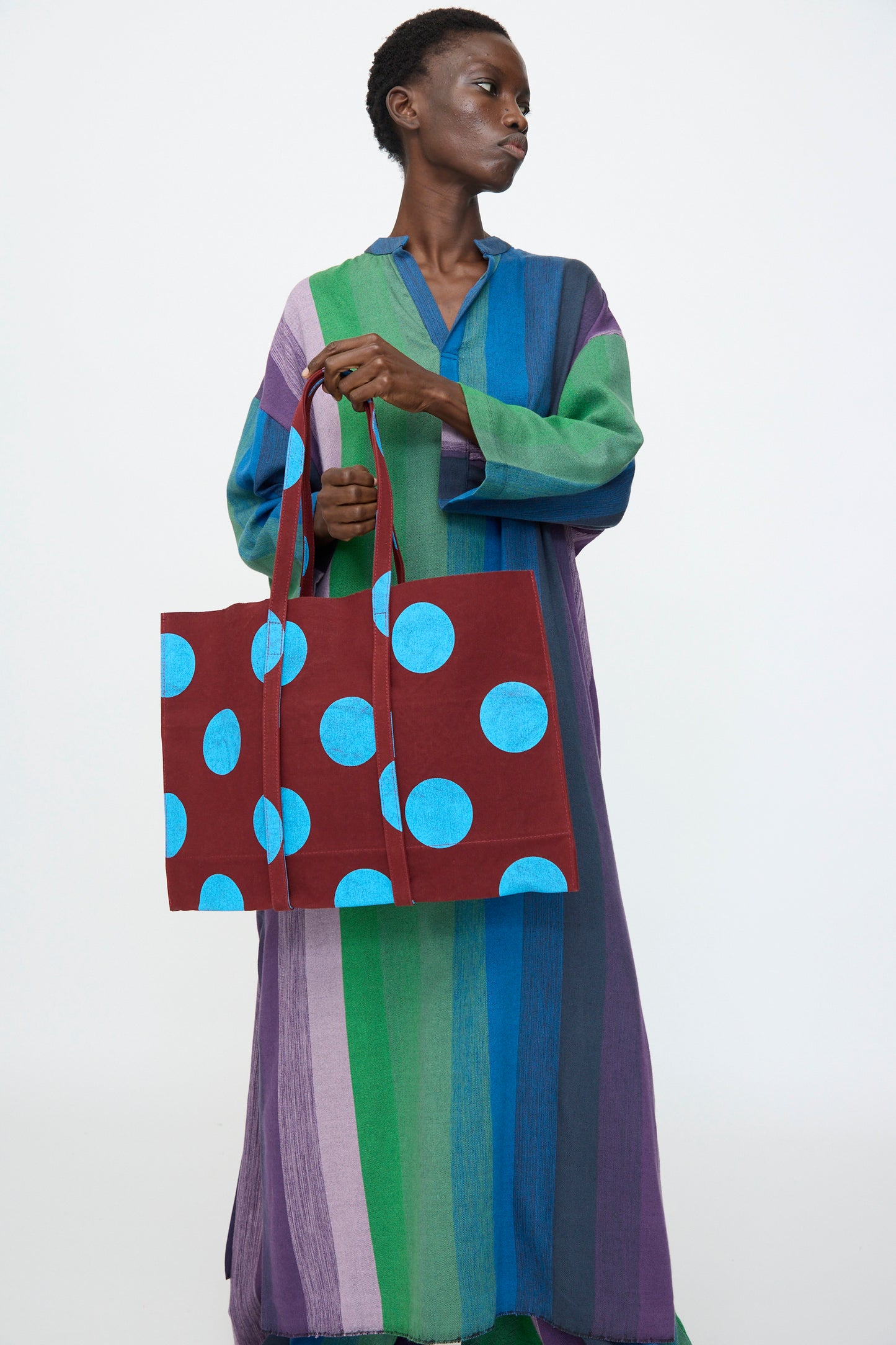 A person in a long, multicolored striped dress holds the Amiacalva Washed Canvas Medium Dot Tote in Sky Blue, made in Japan, against a plain background.